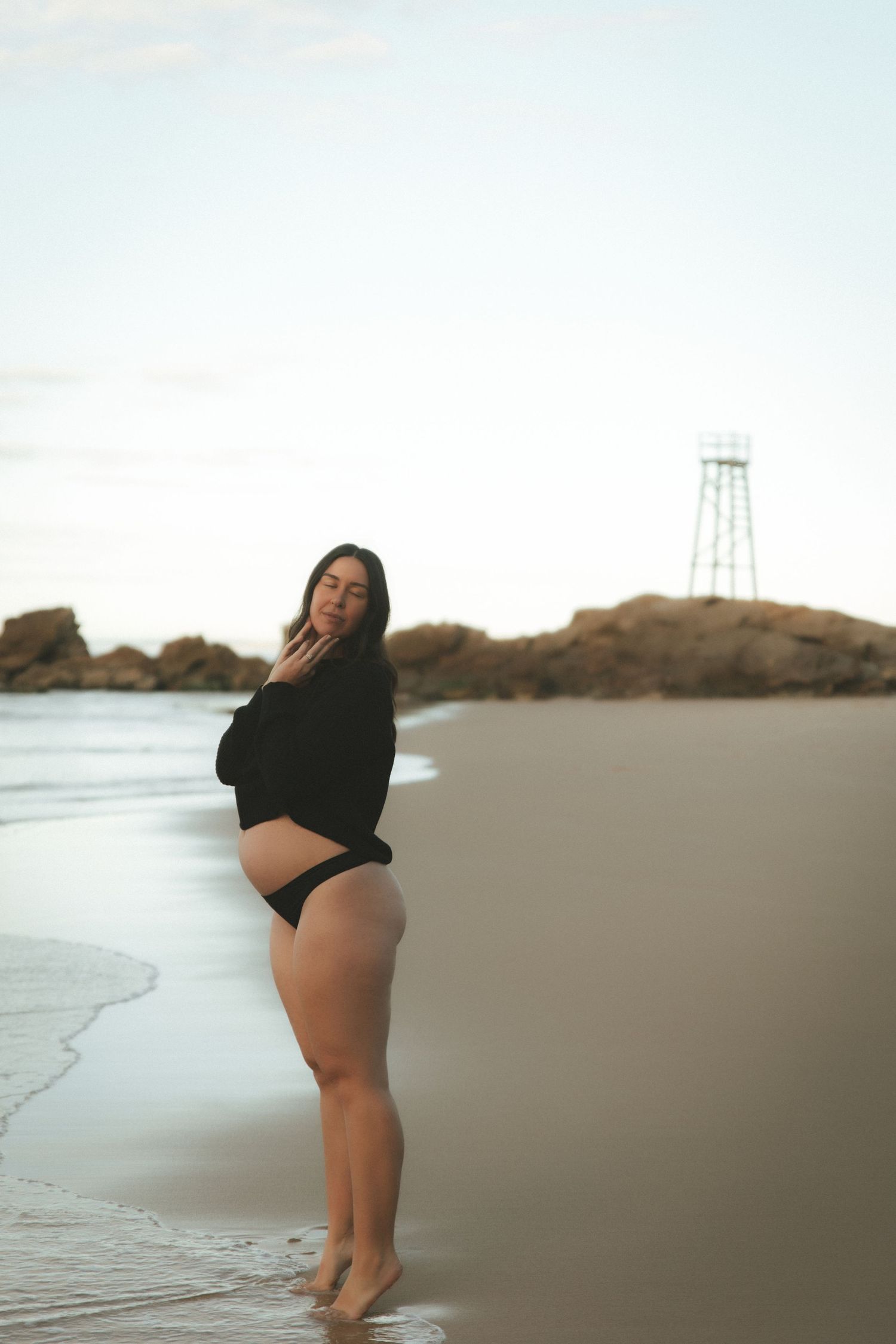 Pregnant woman in black top and bikini bottoms posing on a sandy beach at dusk with rocks in the background.