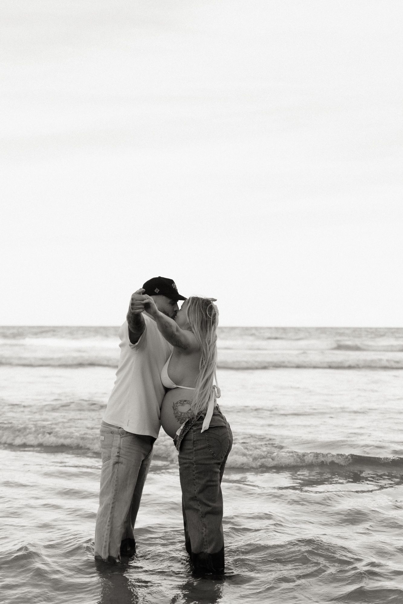Black and white photo of a couple kissing while standing in ocean waves at the beach.