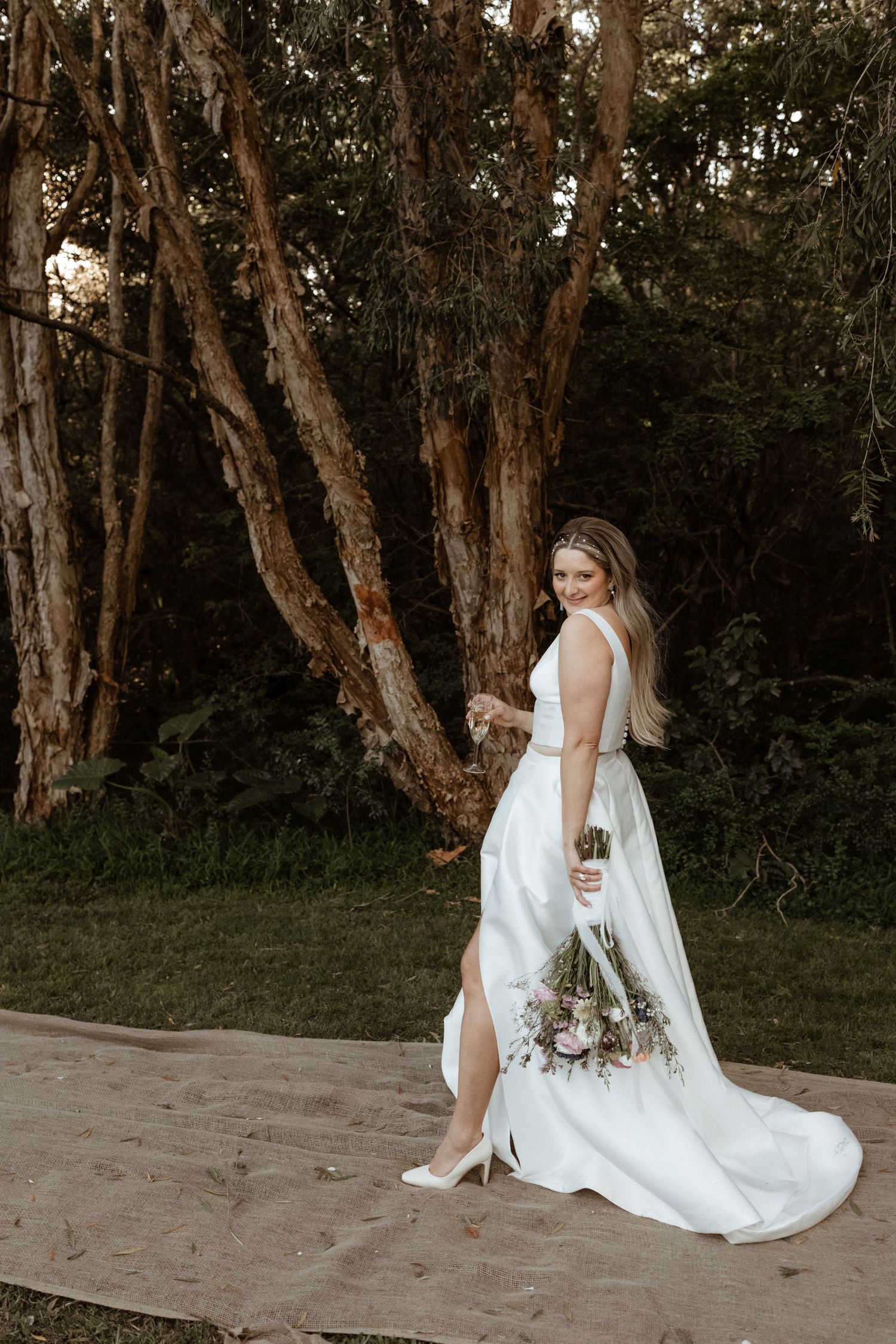Bride in elegant white gown with slit holding a floral bouquet posing outdoors among tall trees.