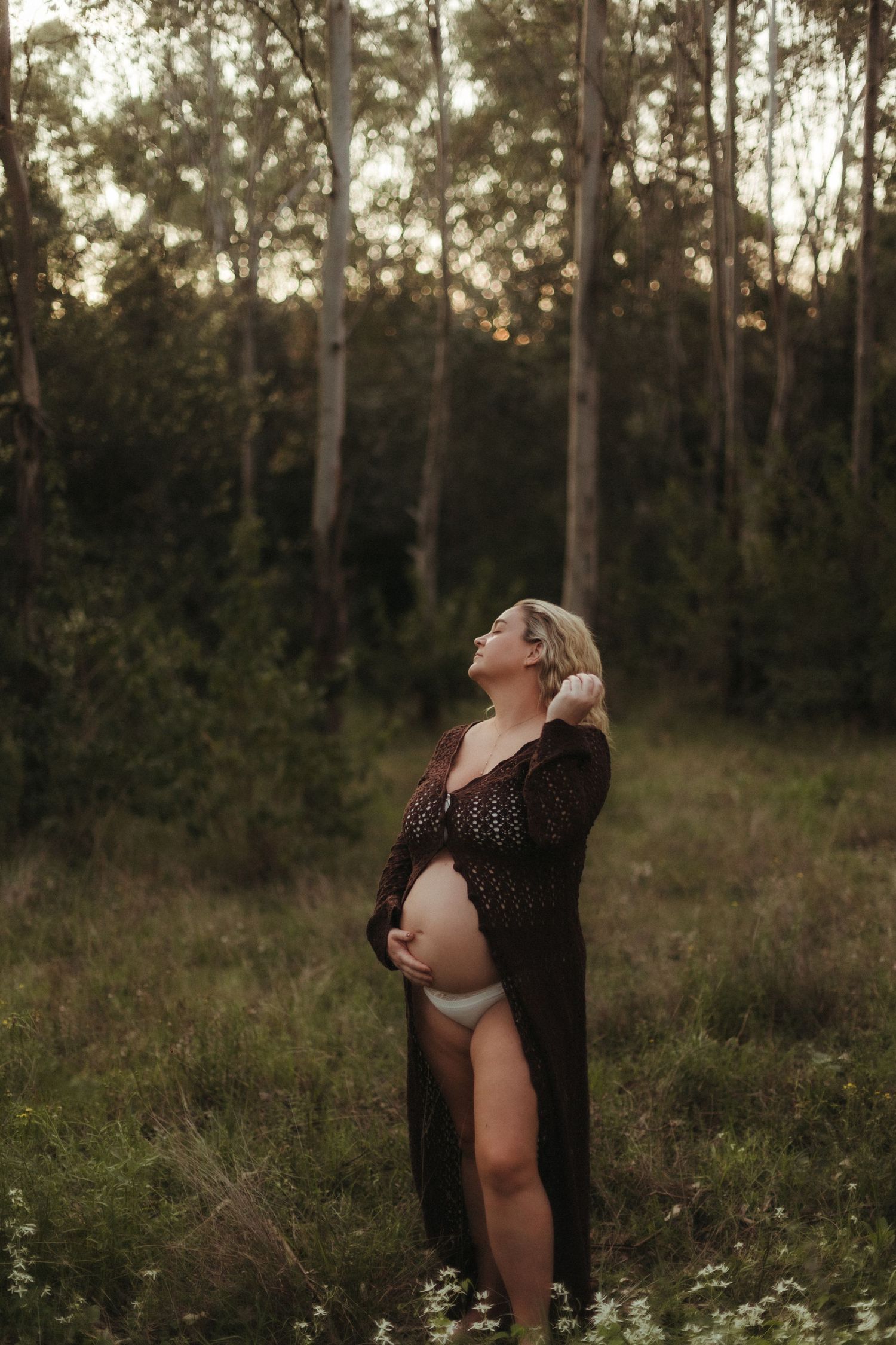 Pregnant woman in dark lace robe standing in forest clearing at dusk.