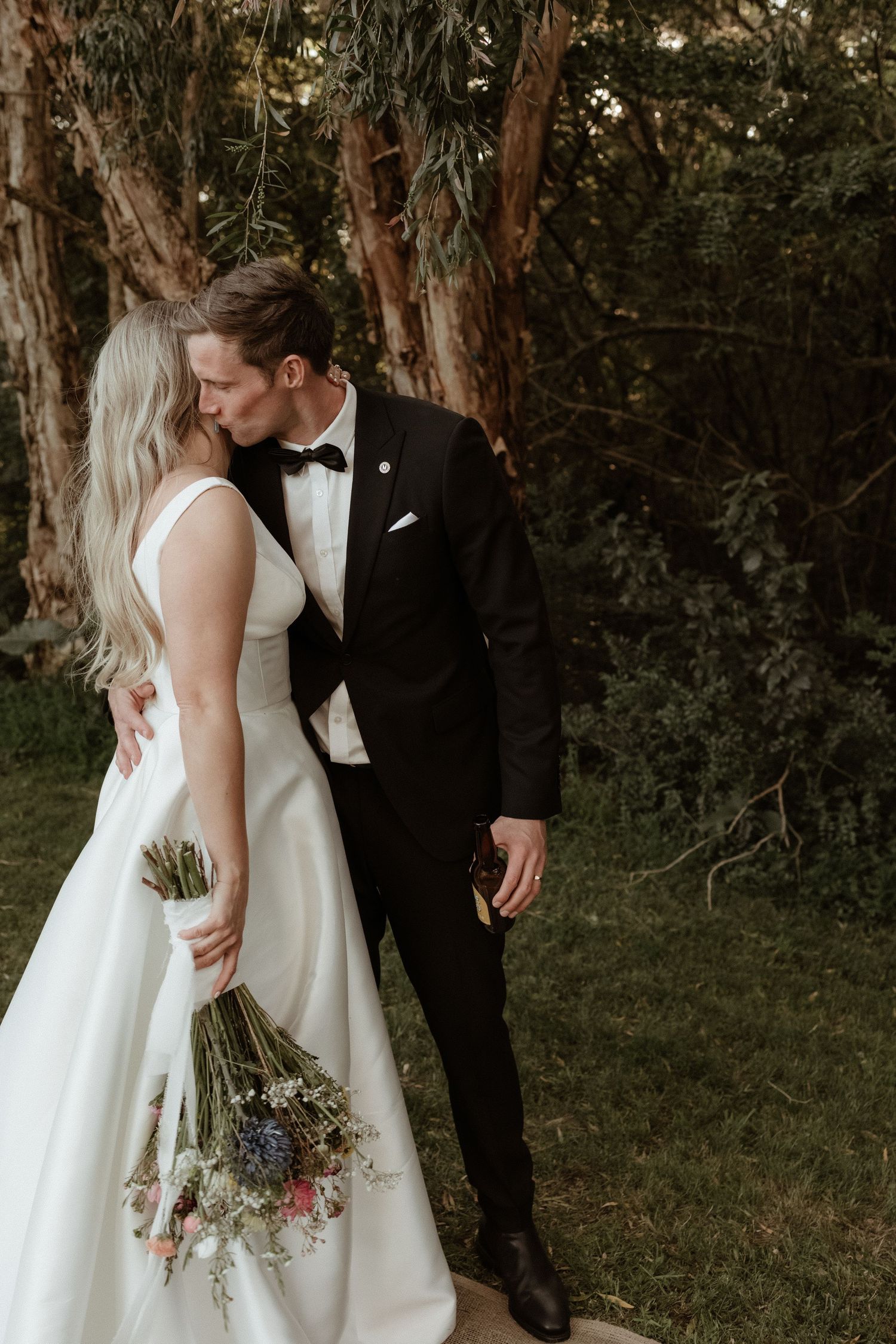 Bride and groom sharing a kiss in a wooded garden, bride in white gown, groom in black tuxedo.