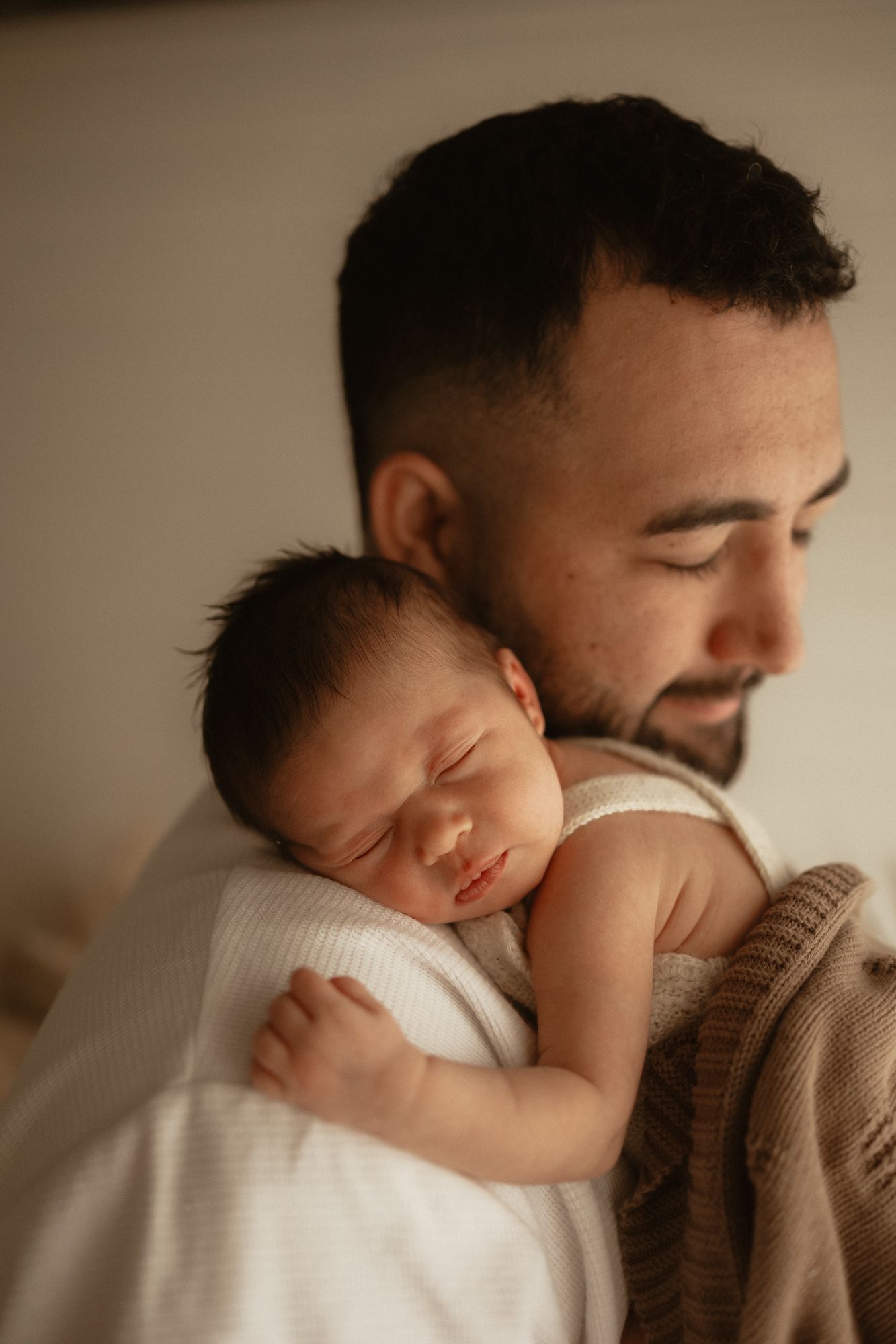 A bearded father tenderly holds his sleeping newborn baby against his chest in a warm, softly lit portrait.