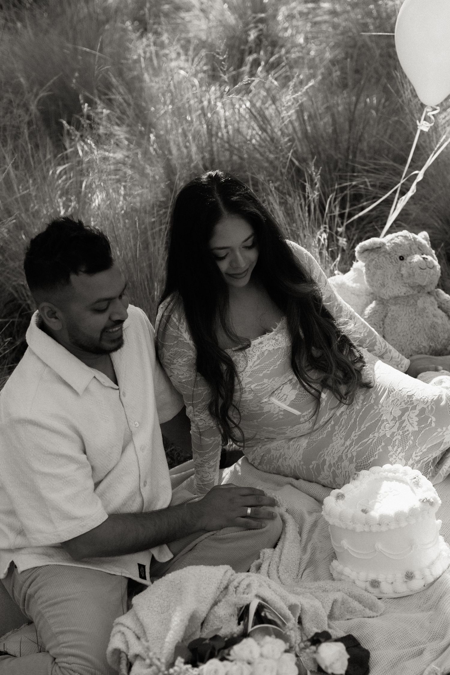 Black and white photo of couple sitting in field with baby shower cake and balloons.