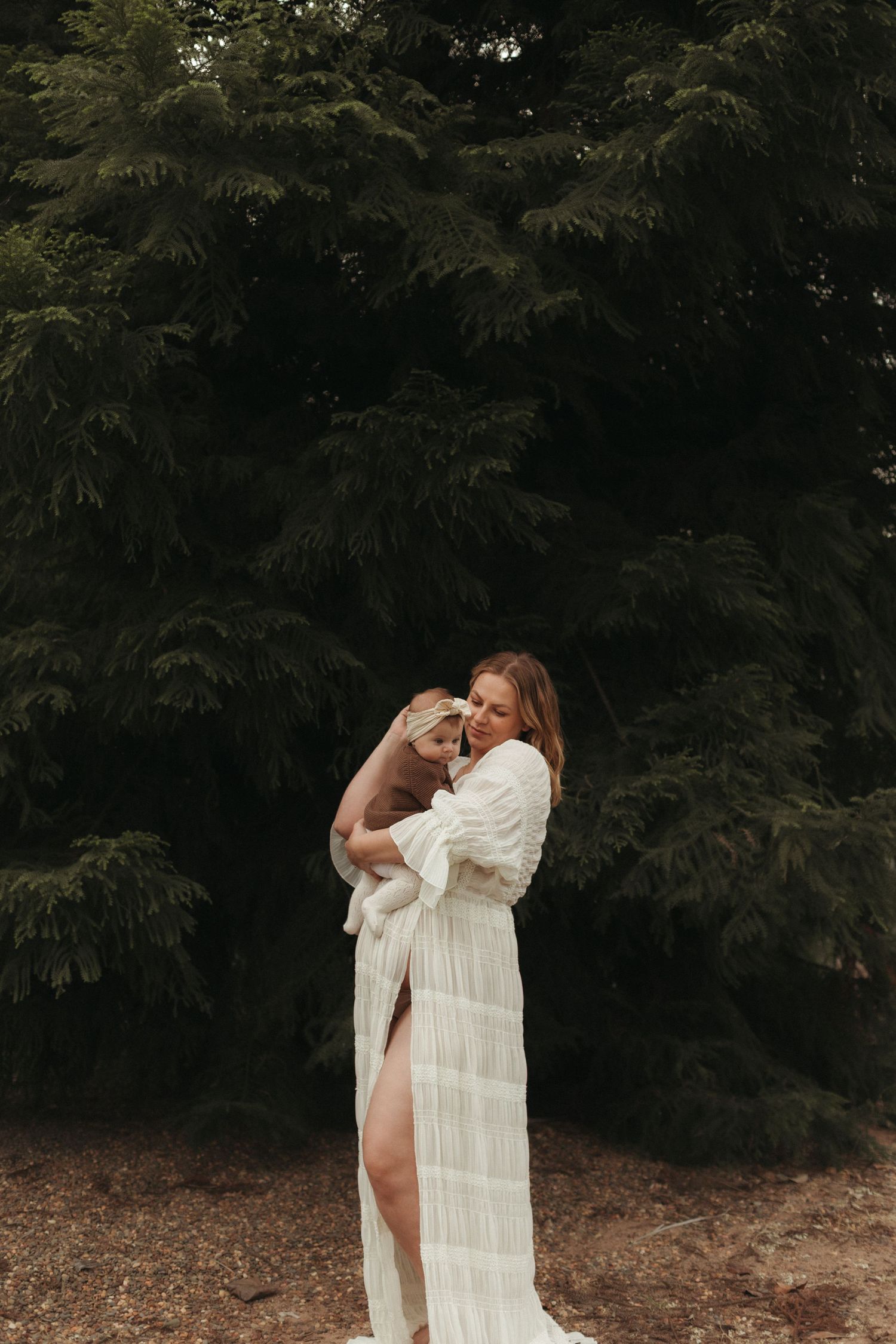 Woman in flowing white dress embracing baby outdoors near tall evergreen trees.