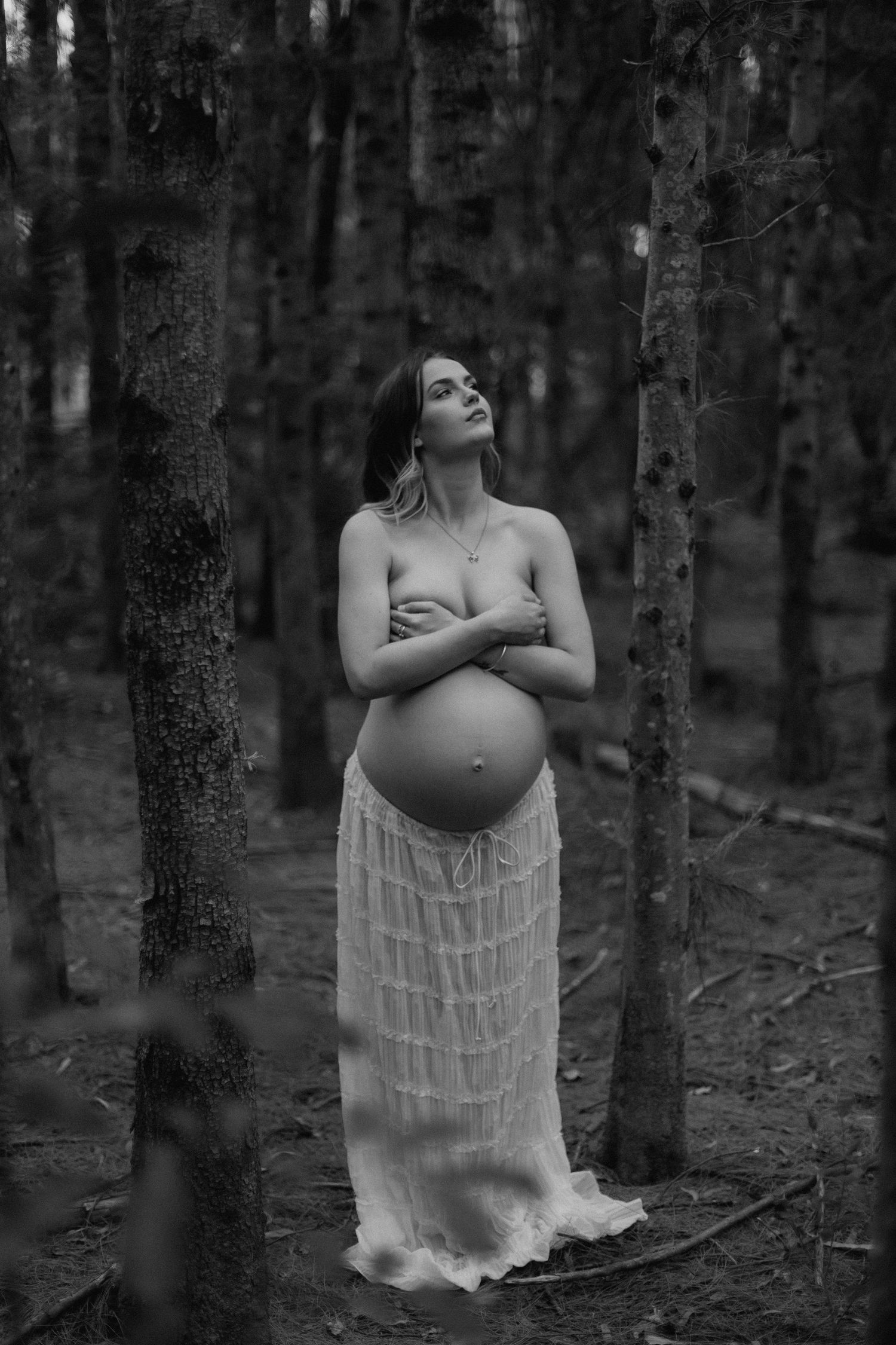 Black and white photo of pregnant woman in lace skirt standing in forest, arms crossed over bare chest.