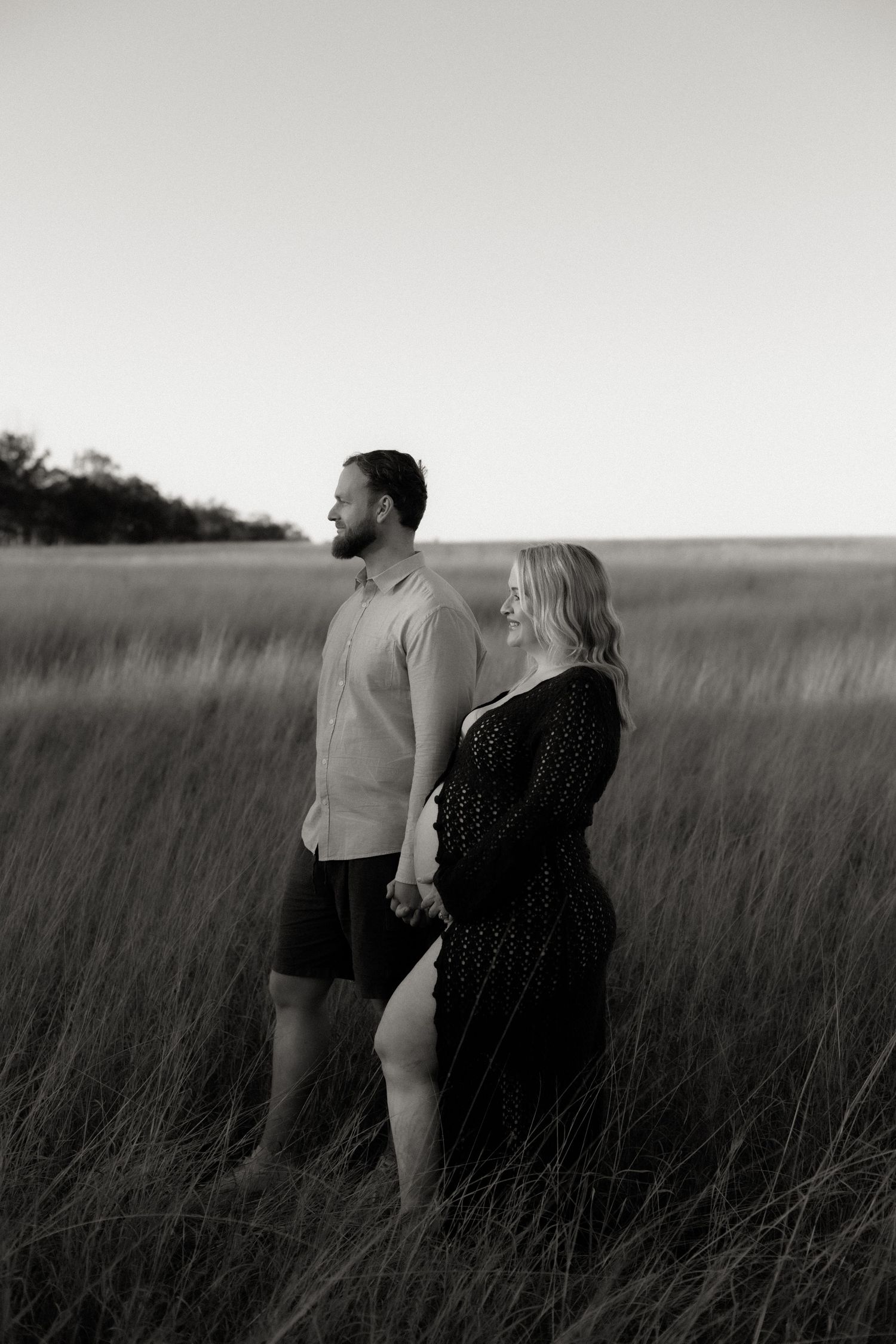 Black and white photo of a pregnant couple holding hands in a golden grass field at sunset.