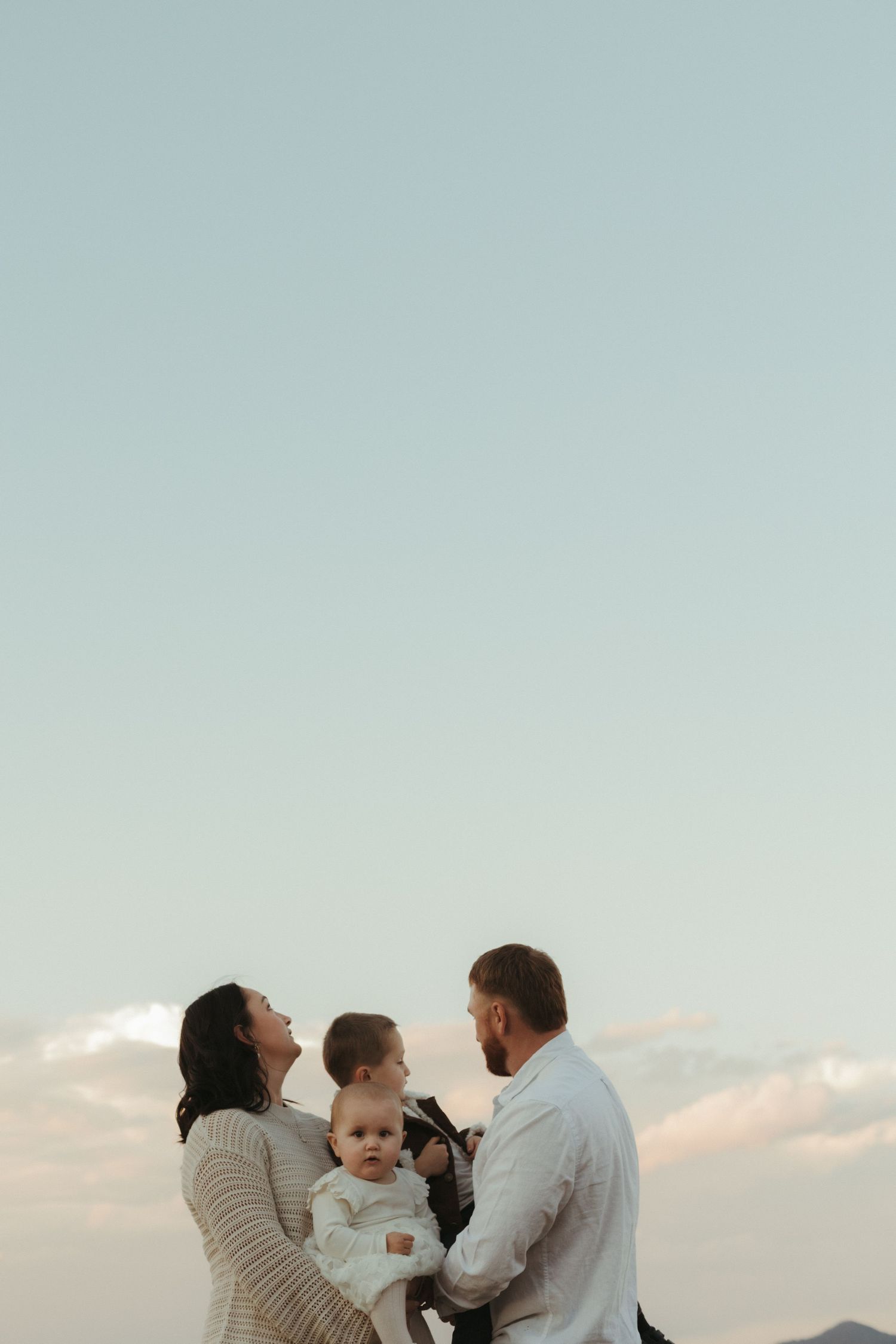 Family with two young children standing together under a soft pastel evening sky outdoors.