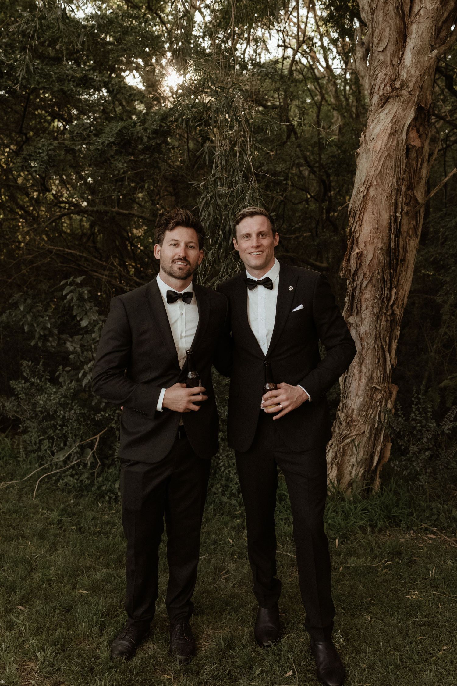 Two grooms in black tuxedos with bow ties smile together outdoors among lush green trees at their wedding.