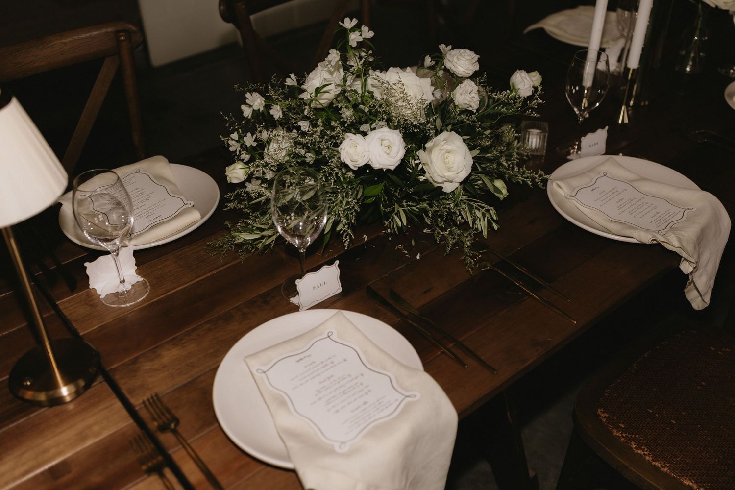 Elegant wedding table setting with white floral centerpiece, white roses, greenery, white plates, and linen napkins on rustic wood table.