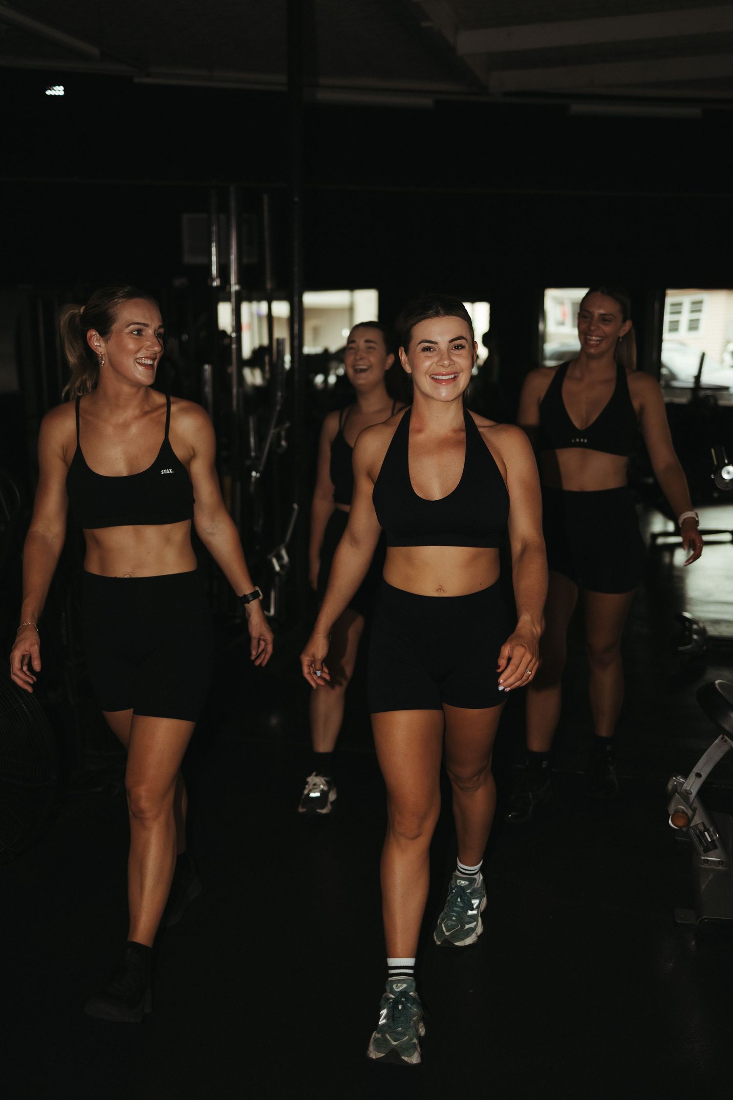 Group of women in matching black activewear walking confidently through dark gym space.