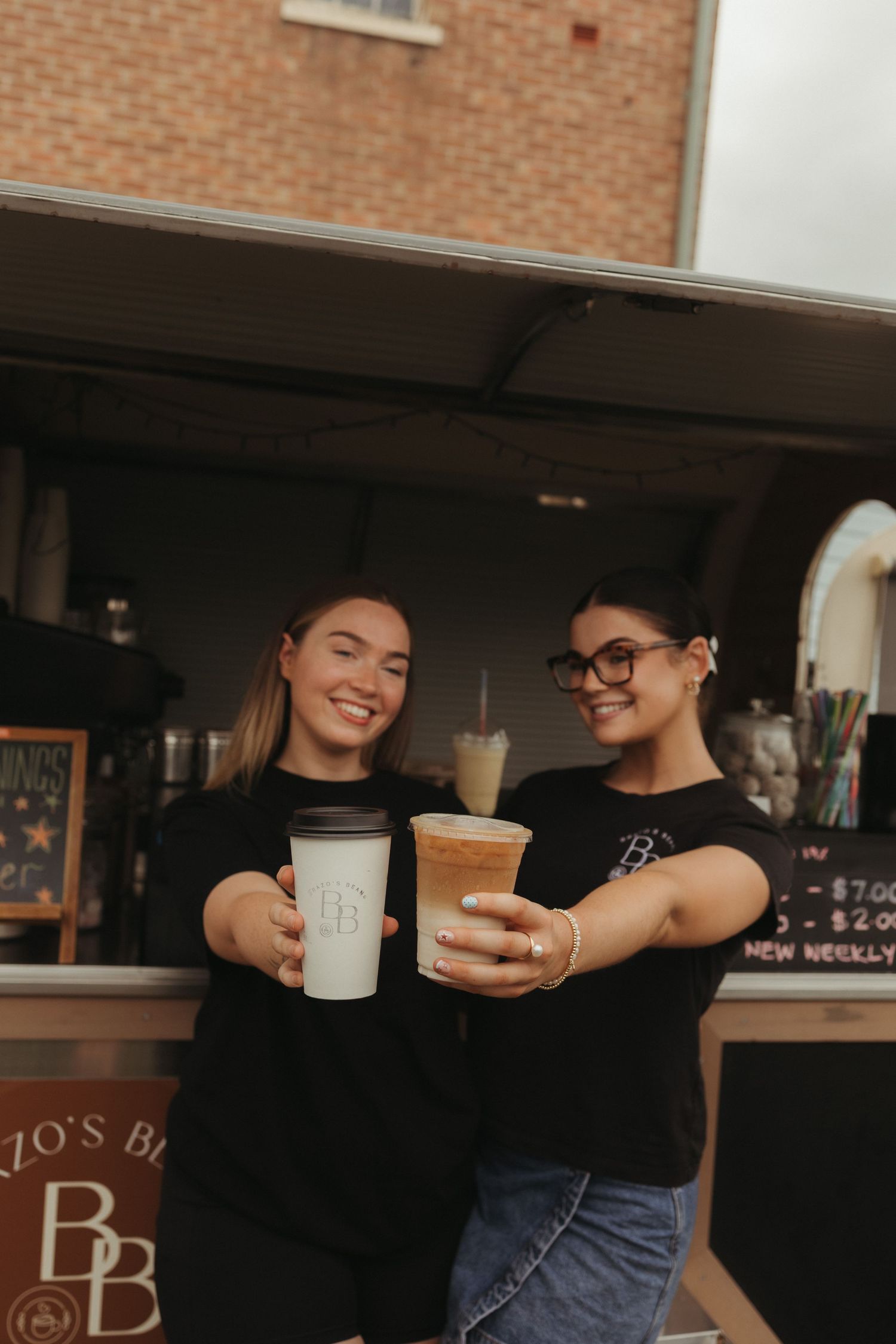 Two smiling women in black BB shirts holding branded coffee cups in front of a food truck.