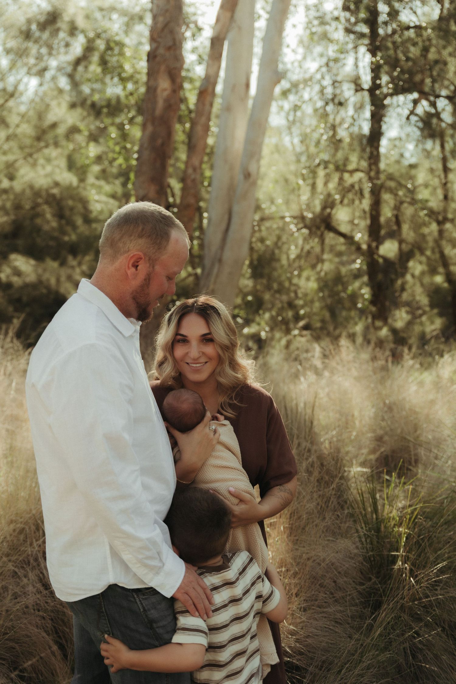 A smiling family with a newborn and young child stand together in a sunlit field surrounded by tall grass.