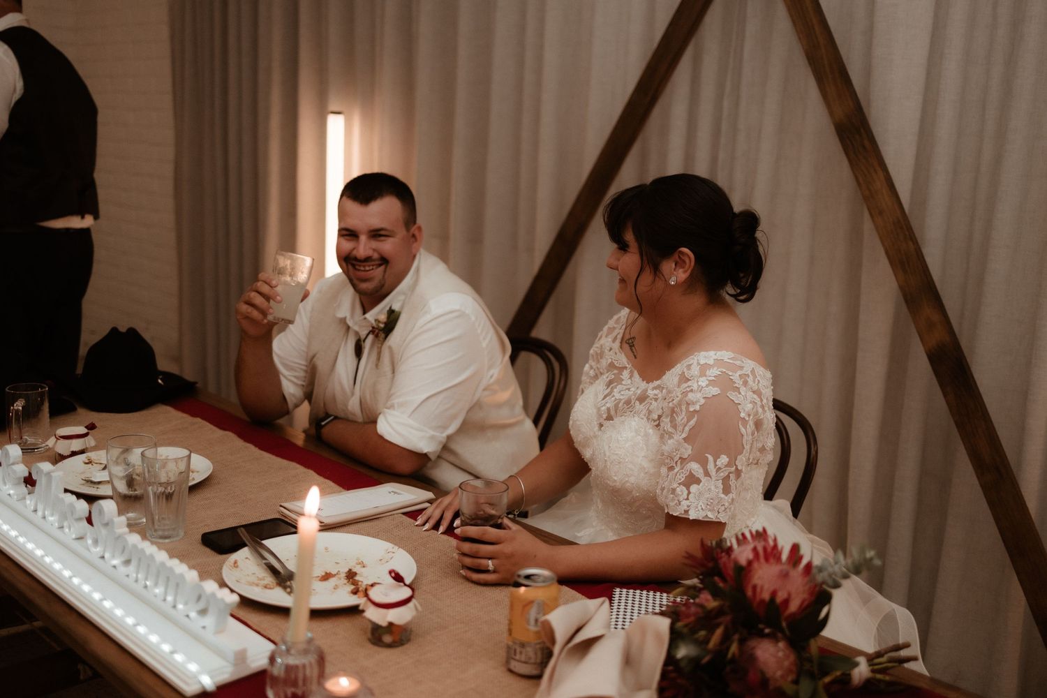 Smiling bride and groom sit together at decorated head table during wedding reception dinner.