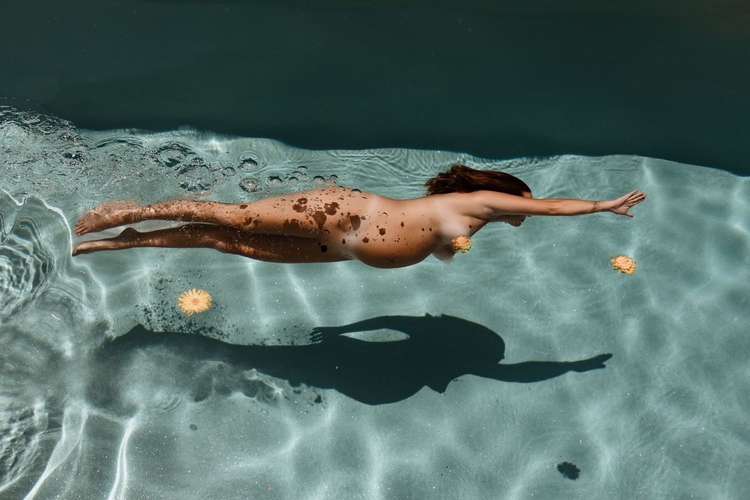 Aerial view of a nude pregnant woman gracefully swimming underwater in a clear turquoise pool.