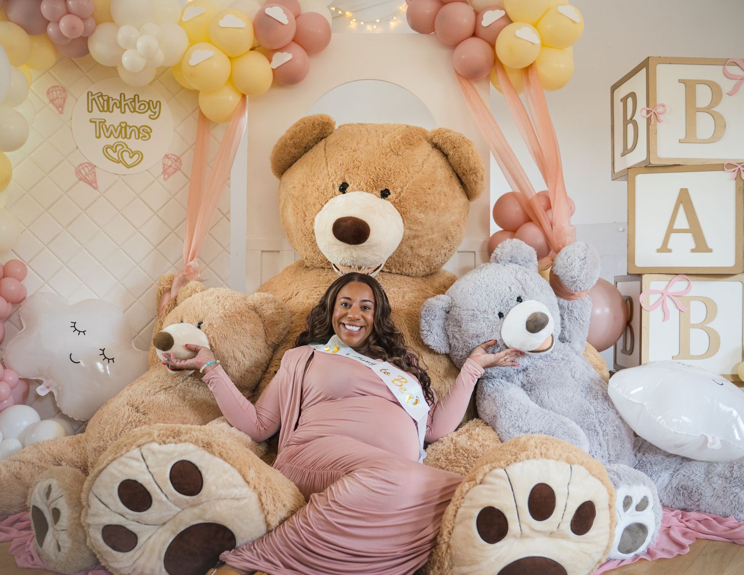 Baby shower decorations with pink and yellow balloons and large teddy bears against white wall.