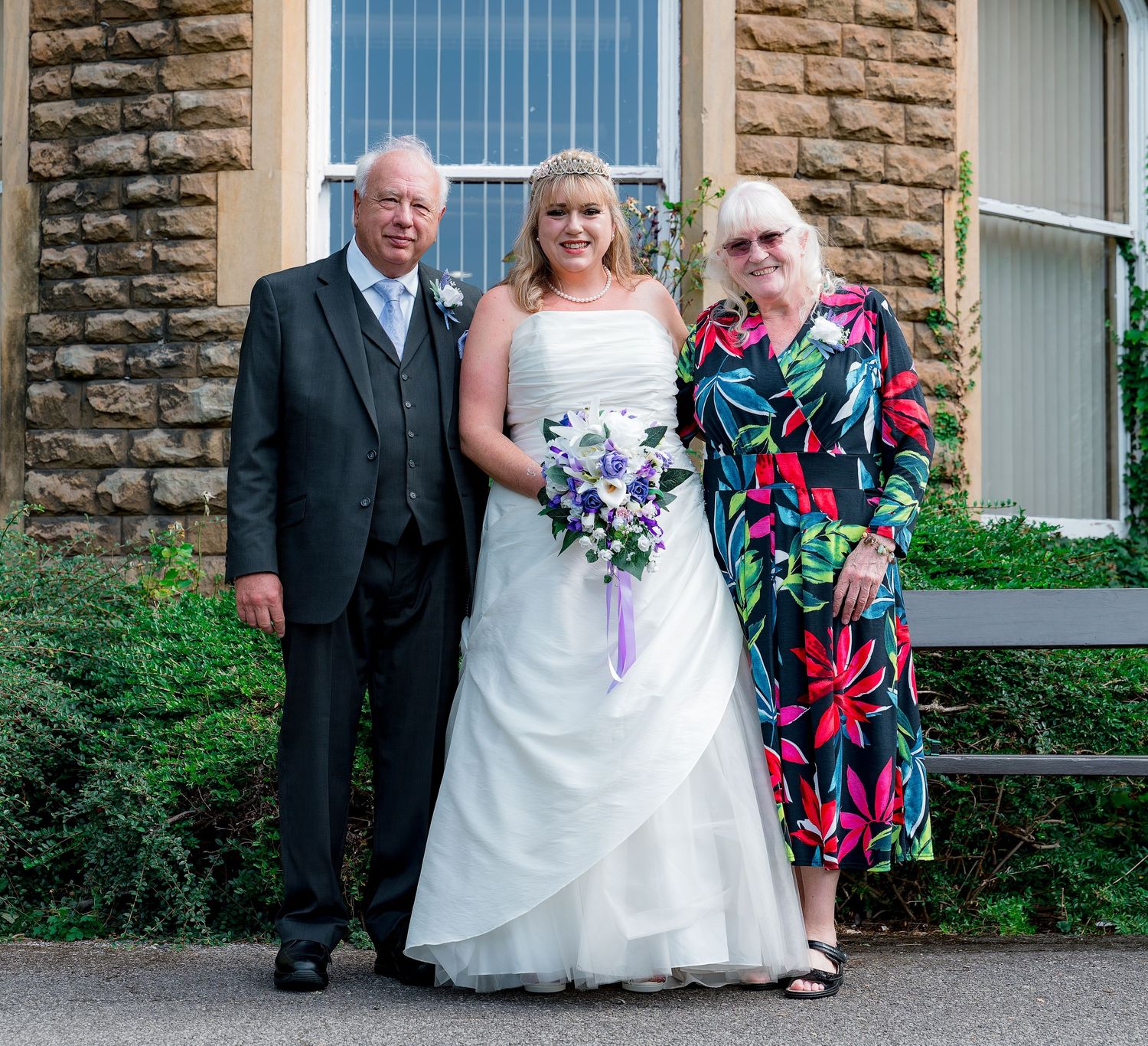 Wedding party poses outside a stone building with arched windows.
