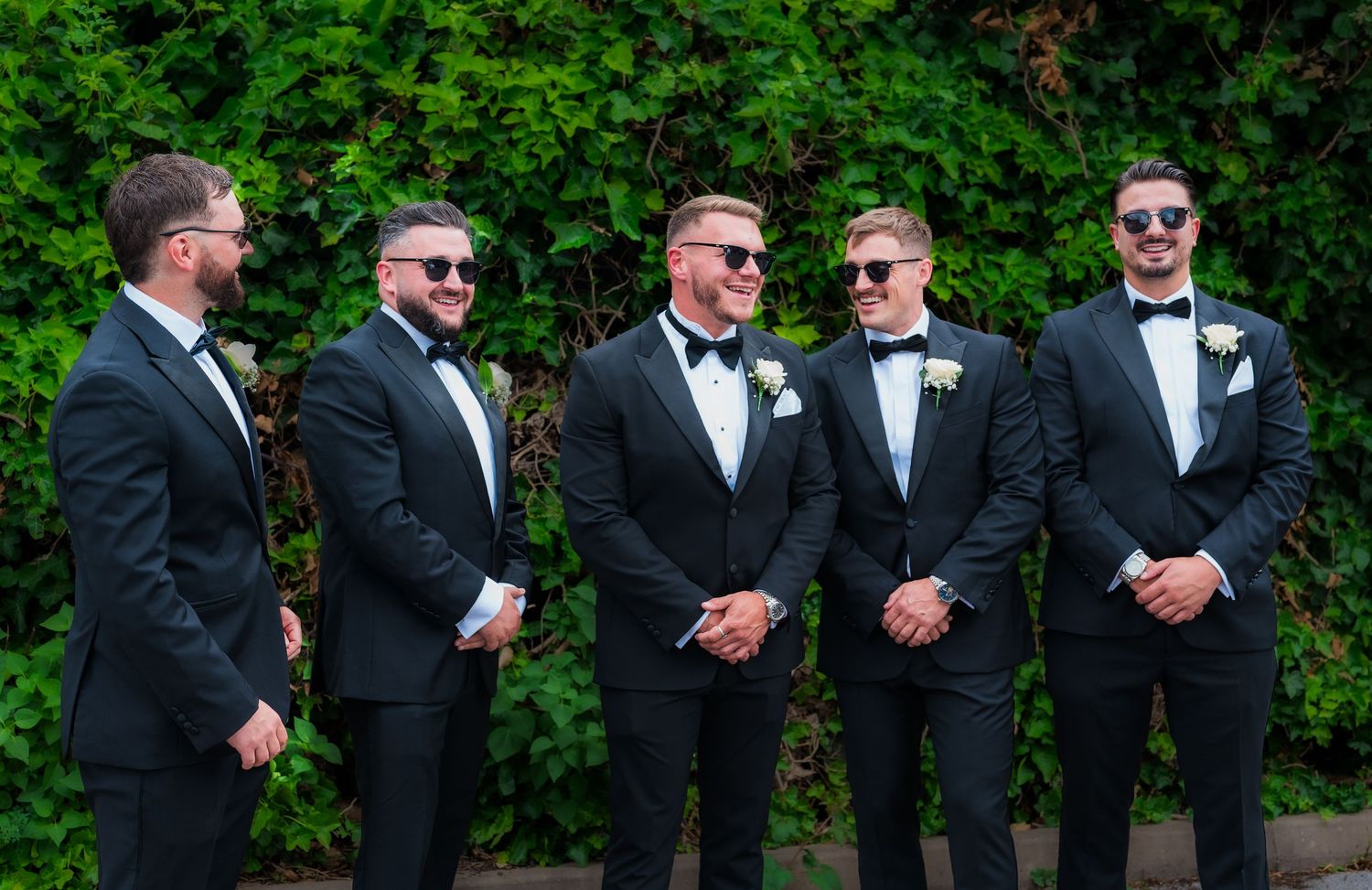 Wedding party in black tuxedos lined up against green hedge backdrop.