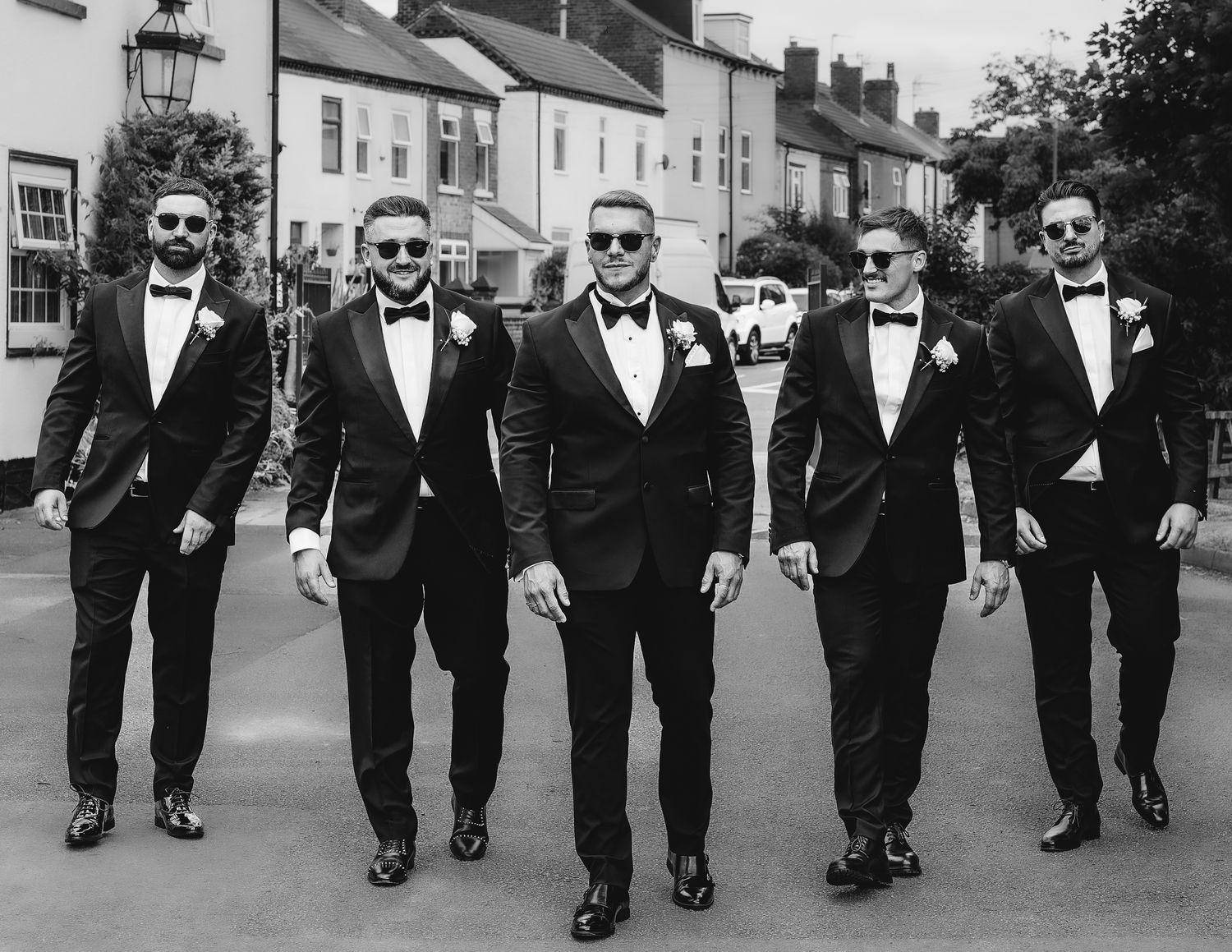 Groomsmen in black tuxedos walking in a fun line formation on a city street.