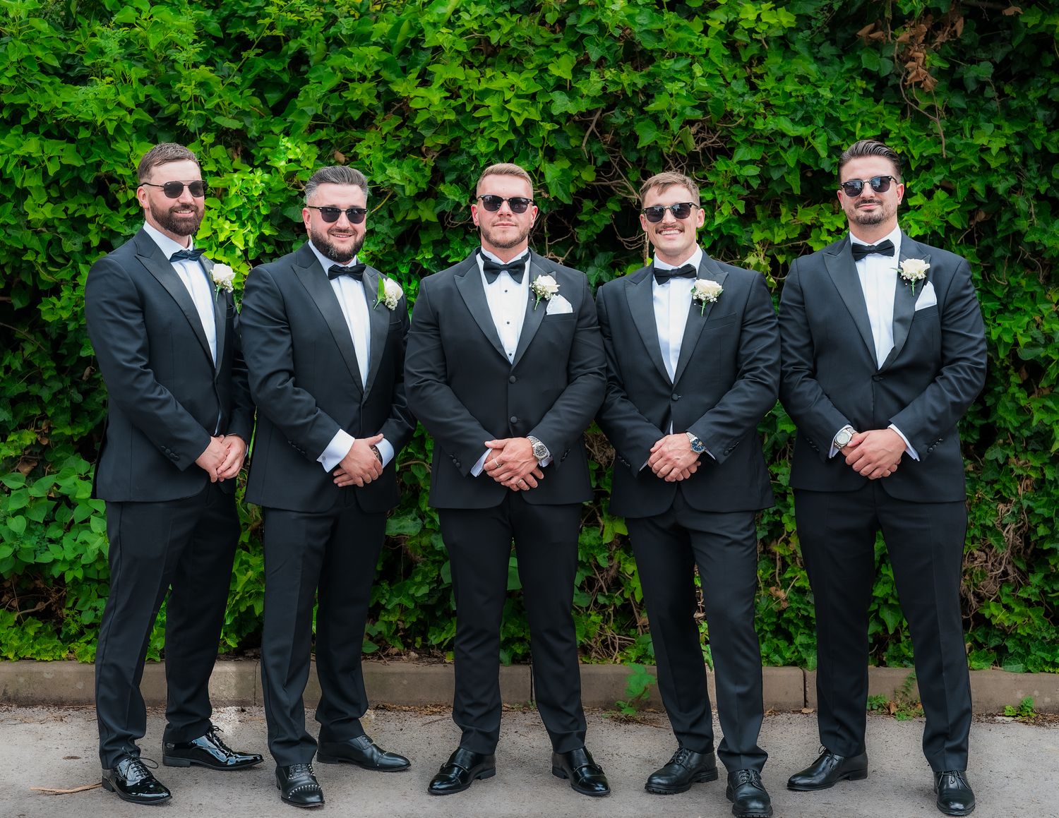 Wedding party members in black suits posing together against lush green garden backdrop.