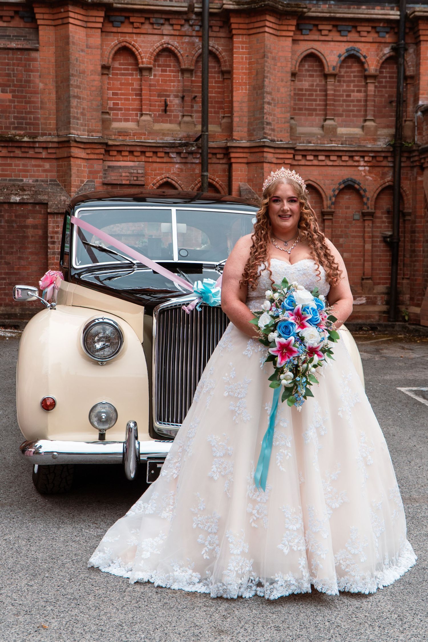 Two people in white wedding dresses stand next to a classic cream-colored Rolls Royce car outside a brick building.