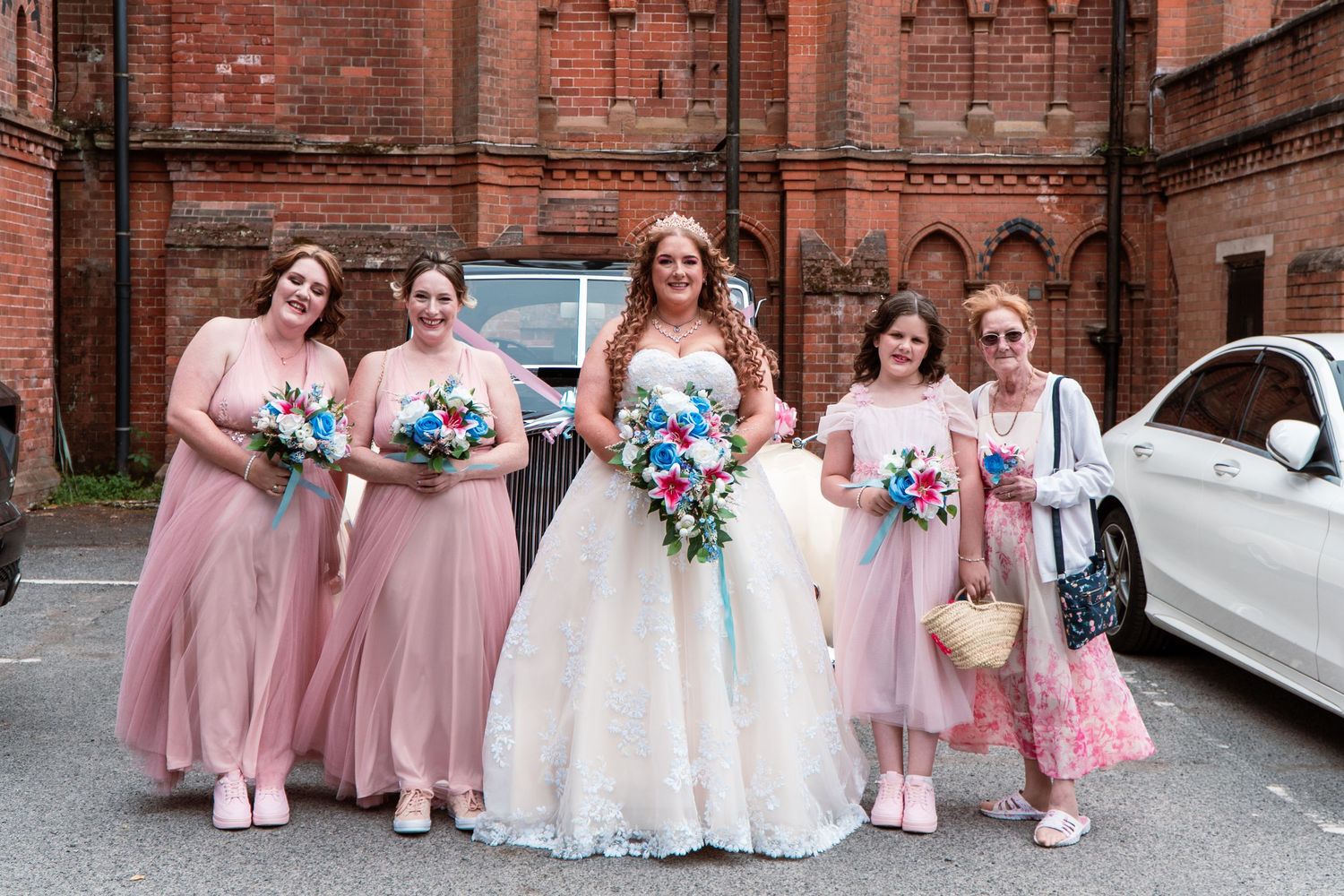 A wedding party poses together outside a brick church wearing pink and white dresses with blue floral bouquets.