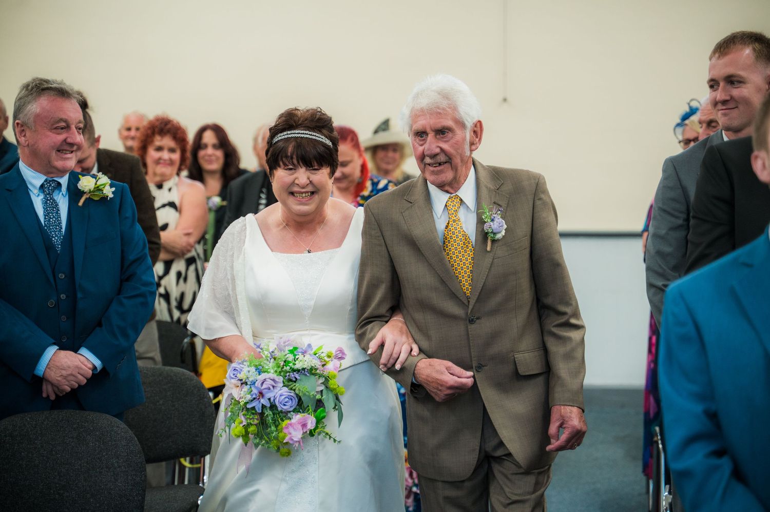 Wedding guests gather outside a venue in formal attire with blue and purple floral bouquets visible.
