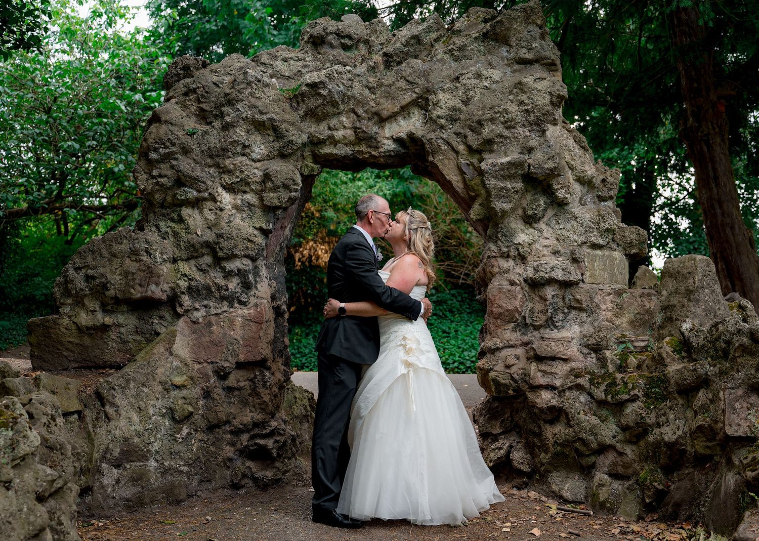 Wedding couple poses against rustic stone wall backdrop in garden setting.