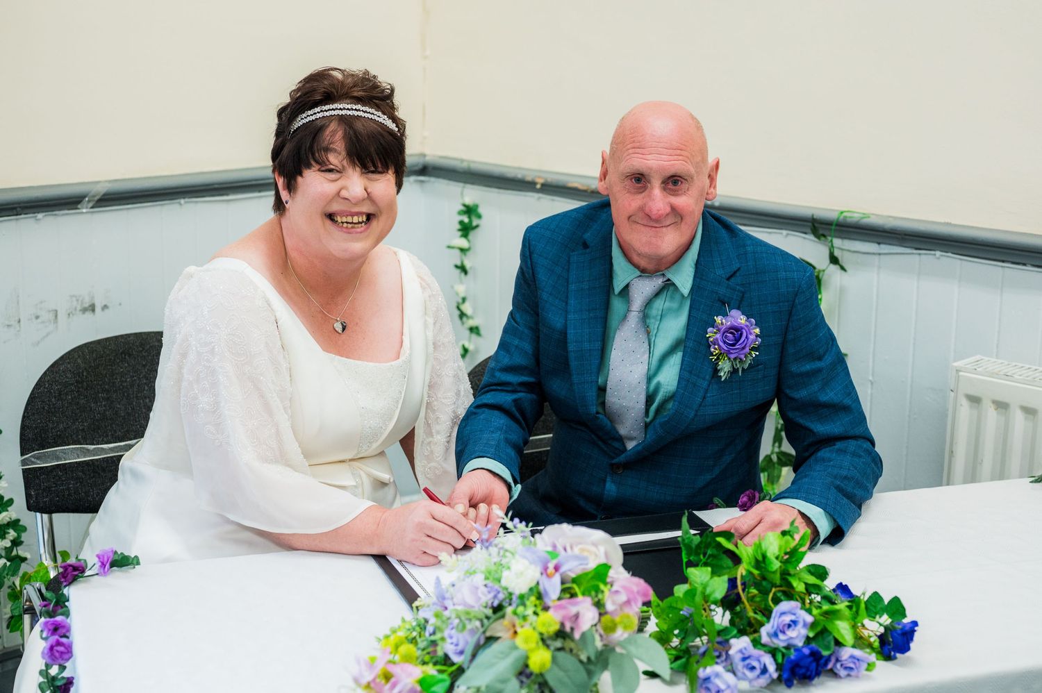 Couple sitting at a table with colorful floral arrangements and paperwork during a signing ceremony.