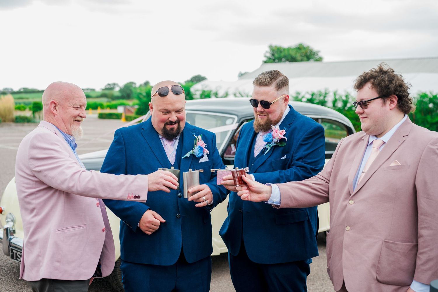A group in formal attire toasting with drinks outside near a parked car on a sunny day.