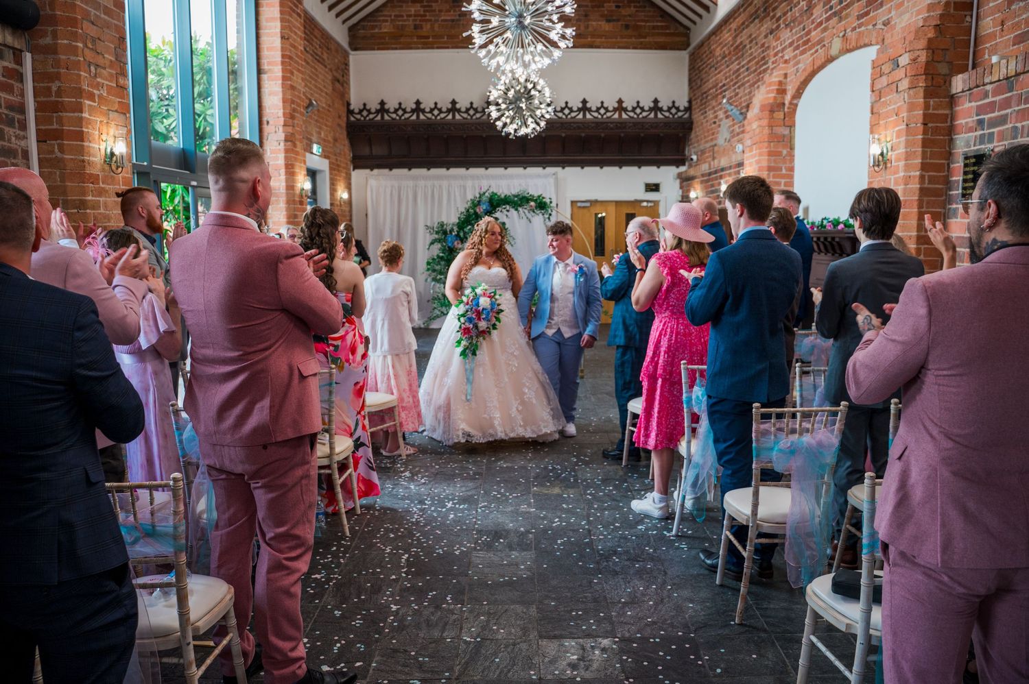 Wedding guests gather in a rustic brick venue with exposed beams and elegant chandeliers during a ceremony.