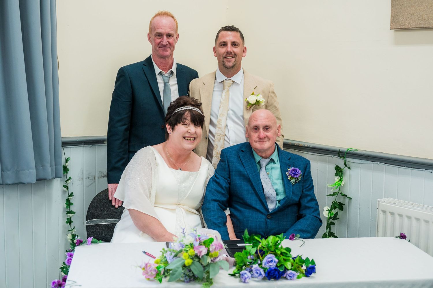 Wedding couple seated at a table with purple and pink floral decorations during document signing.