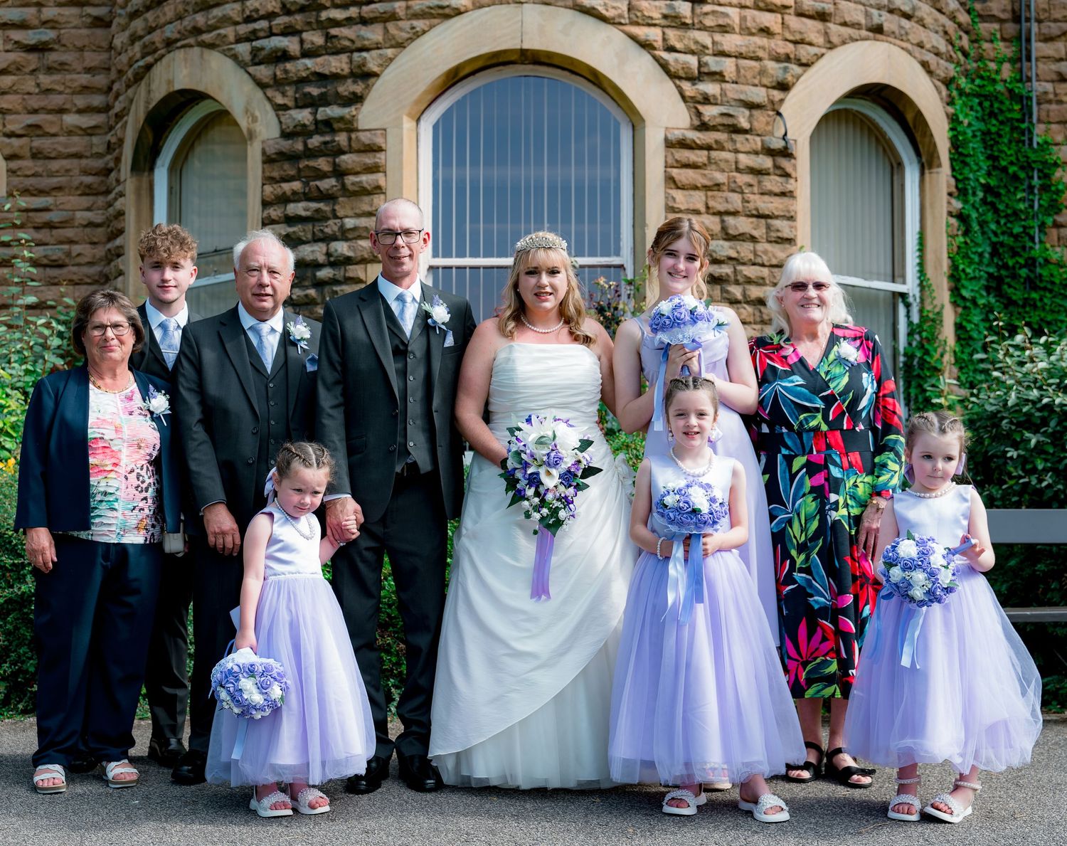 Wedding party with flower girls in purple dresses posed by stone building.
