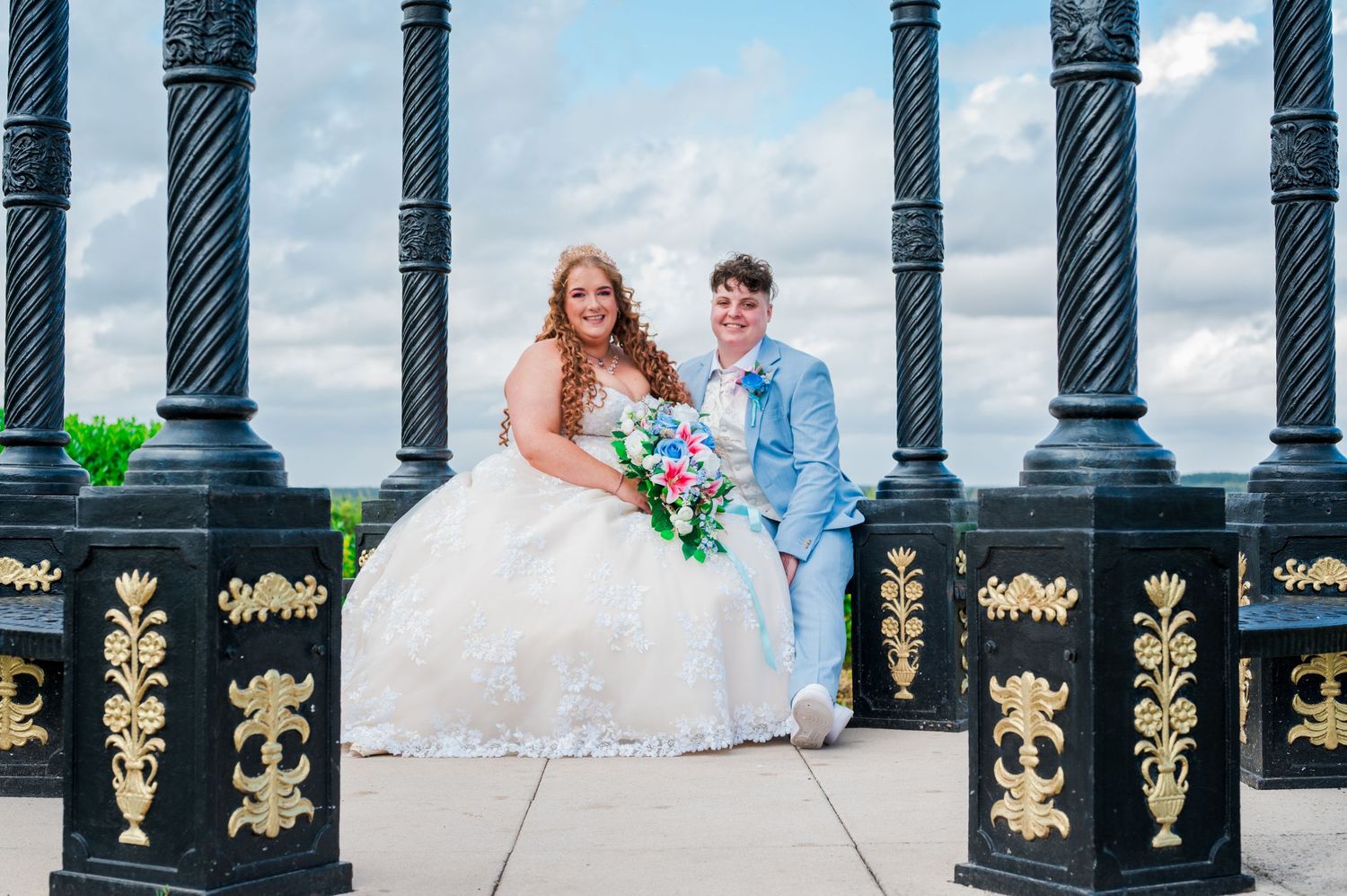 A couple in wedding attire sits on a bench with ornate black and gold iron railings in the background.