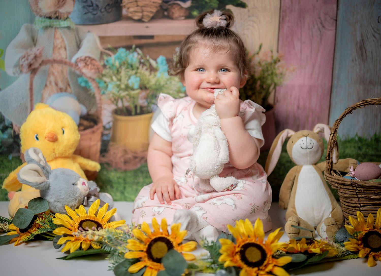 Baby sitting among plush toys and sunflowers in rustic setting.