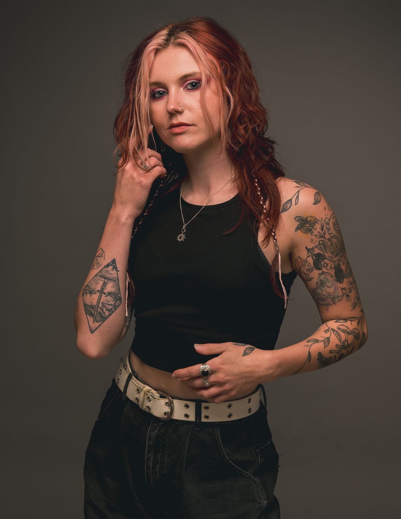 Woman with red hair and tattoos posing in black outfit against gray backdrop.