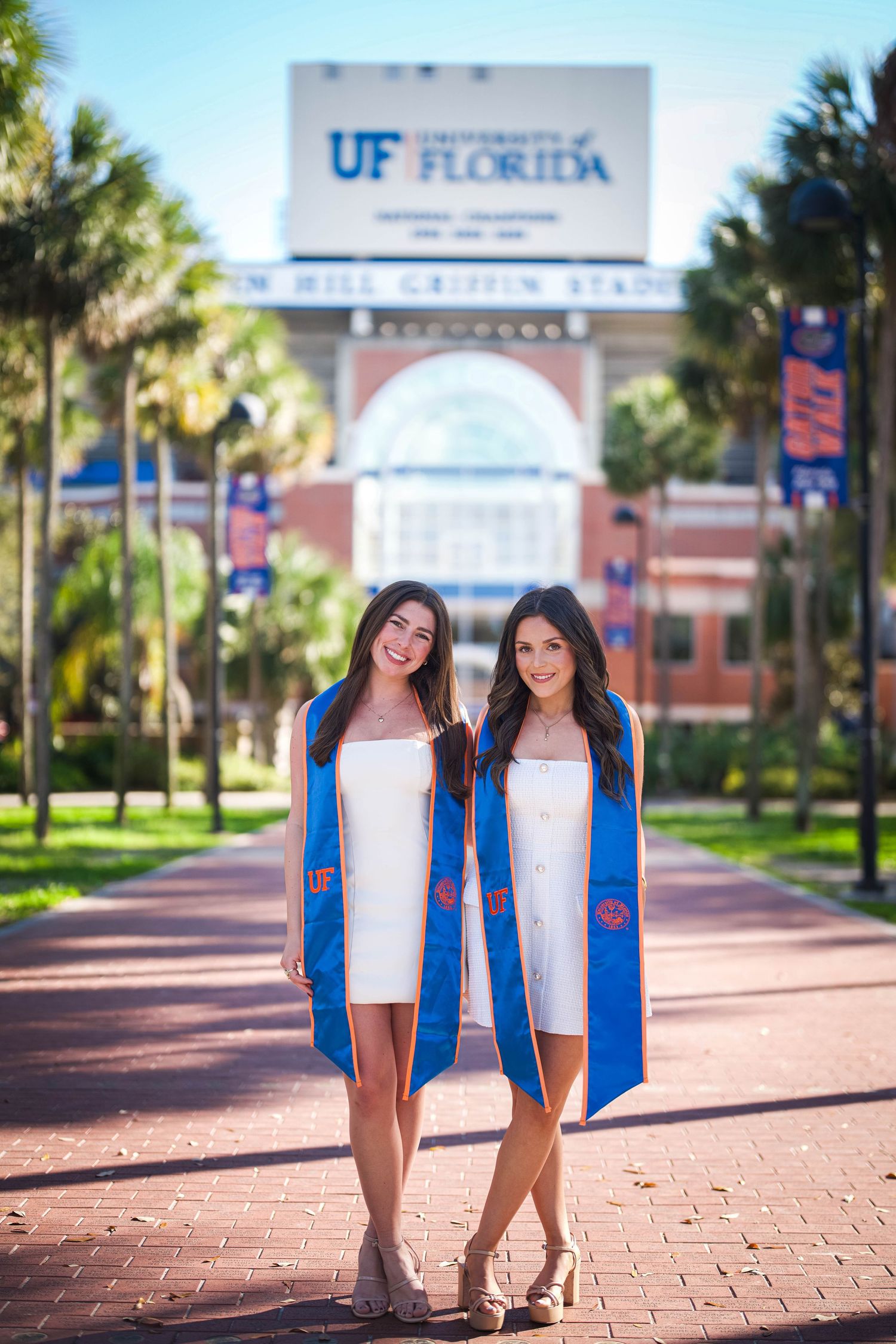 Two graduates in blue and orange stoles pose on a brick pathway in front of the University of Florida stadium called Gator Walk.