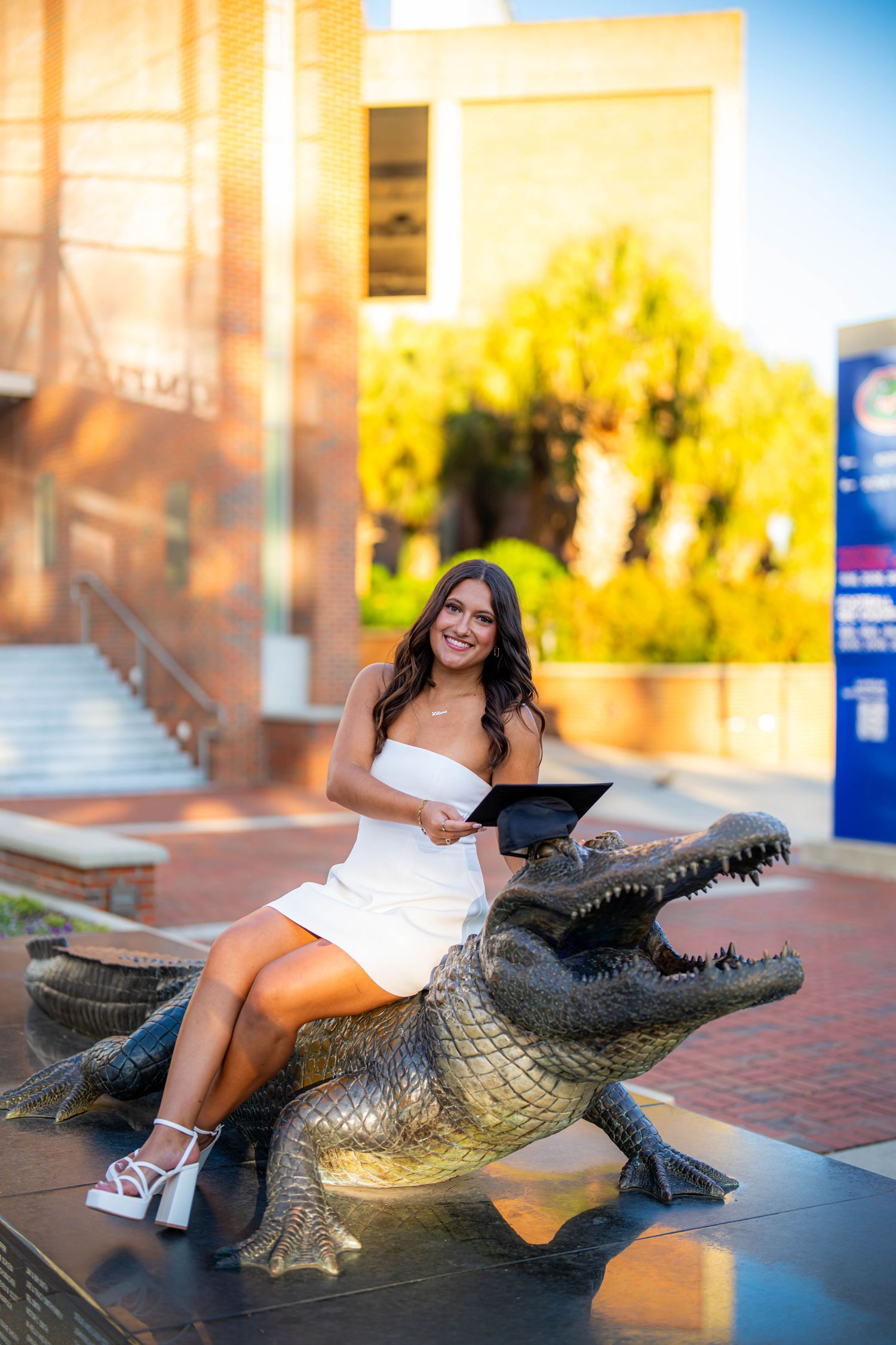 UF Graduate in a white dress poses on the UF Bull Gator Statue on a college campus during golden hour.