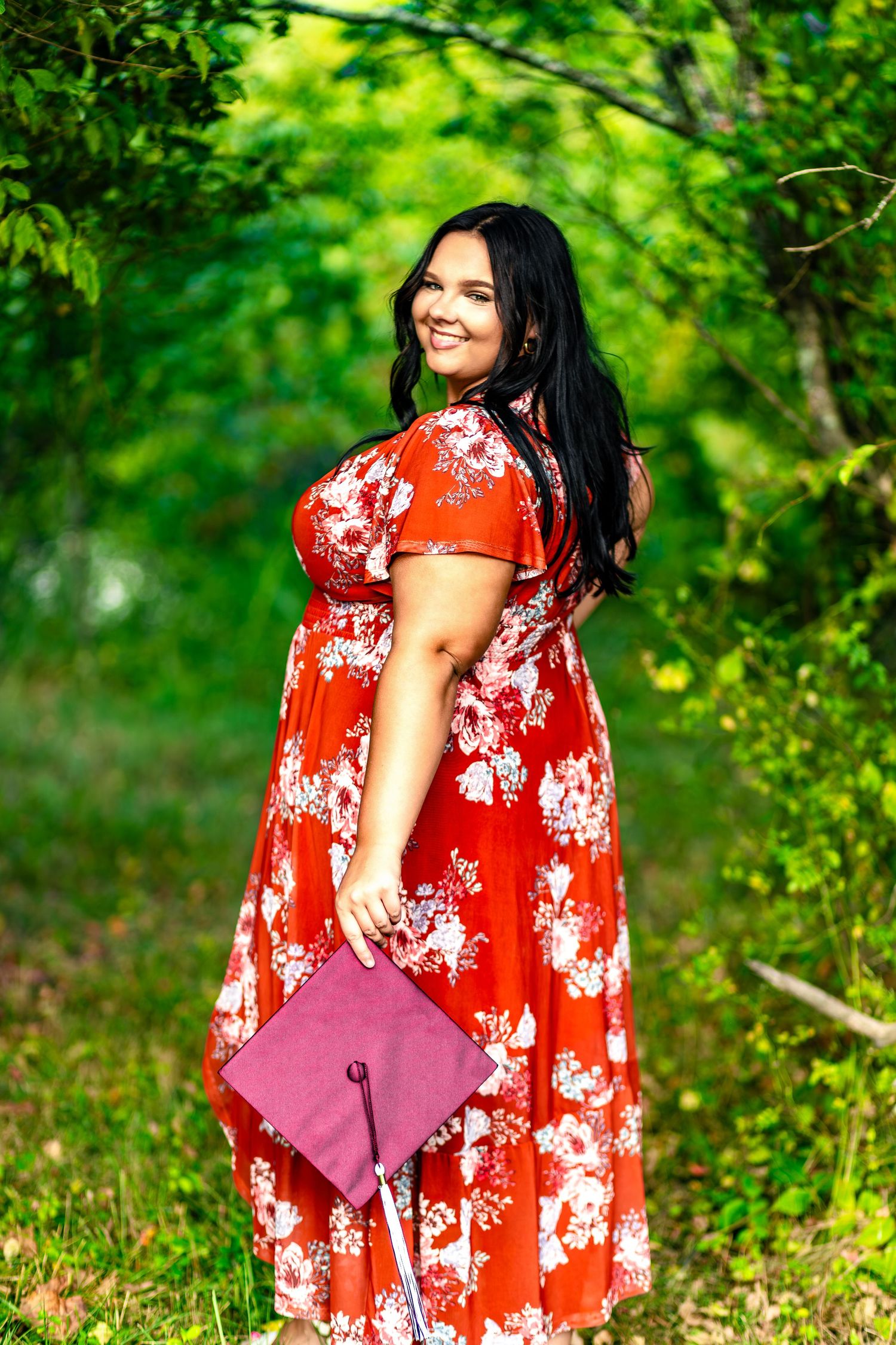 Graduate celebrates graduation in a bright red floral dress among lush green trees holding a burgundy diploma case.