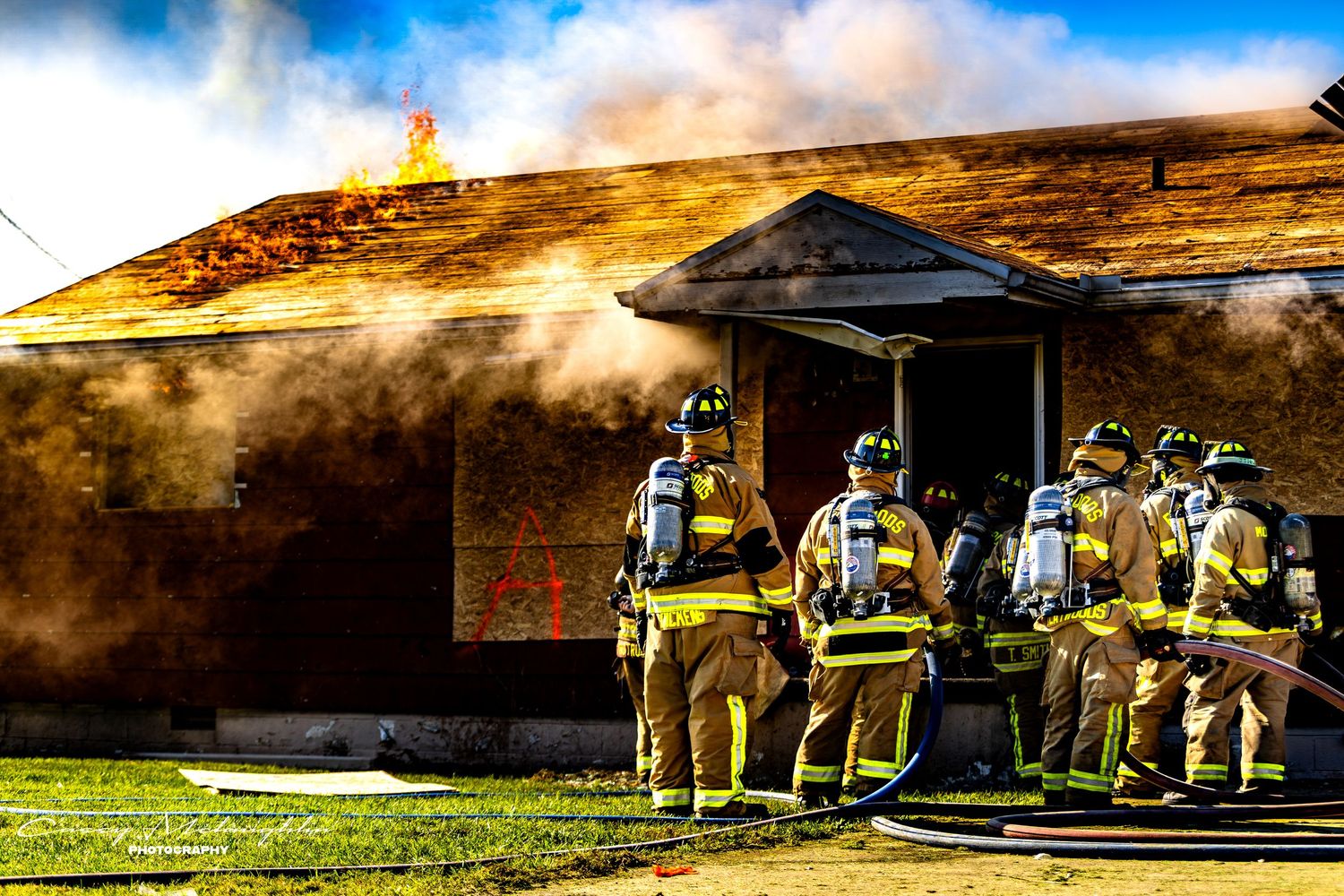 Firefighters in protective gear stand near emergency vehicles during a training exercise at a facility with smoke billowing.