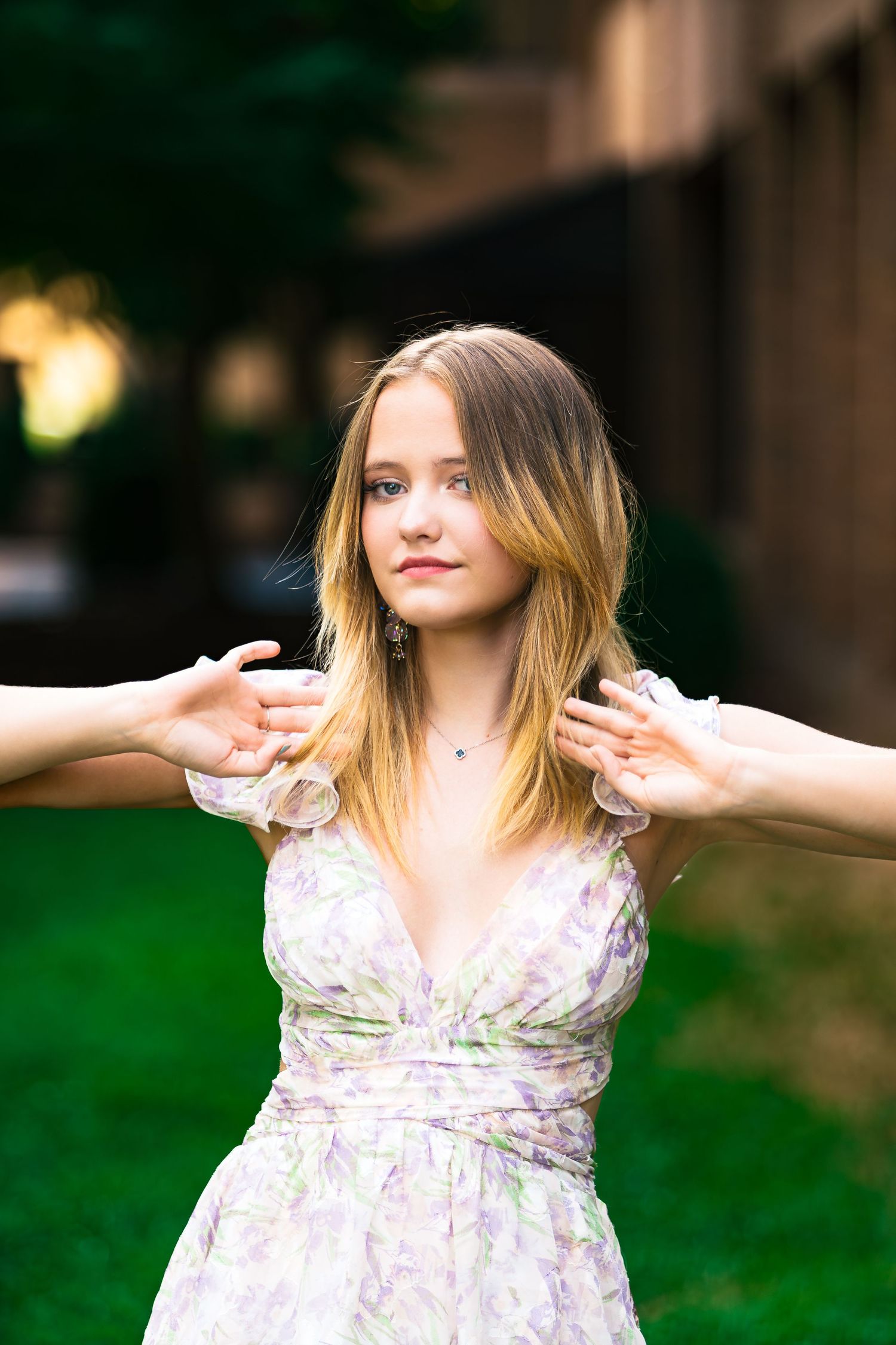 A person in a floral summer dress stands outdoors with arms outstretched as hands reach in from both sides.