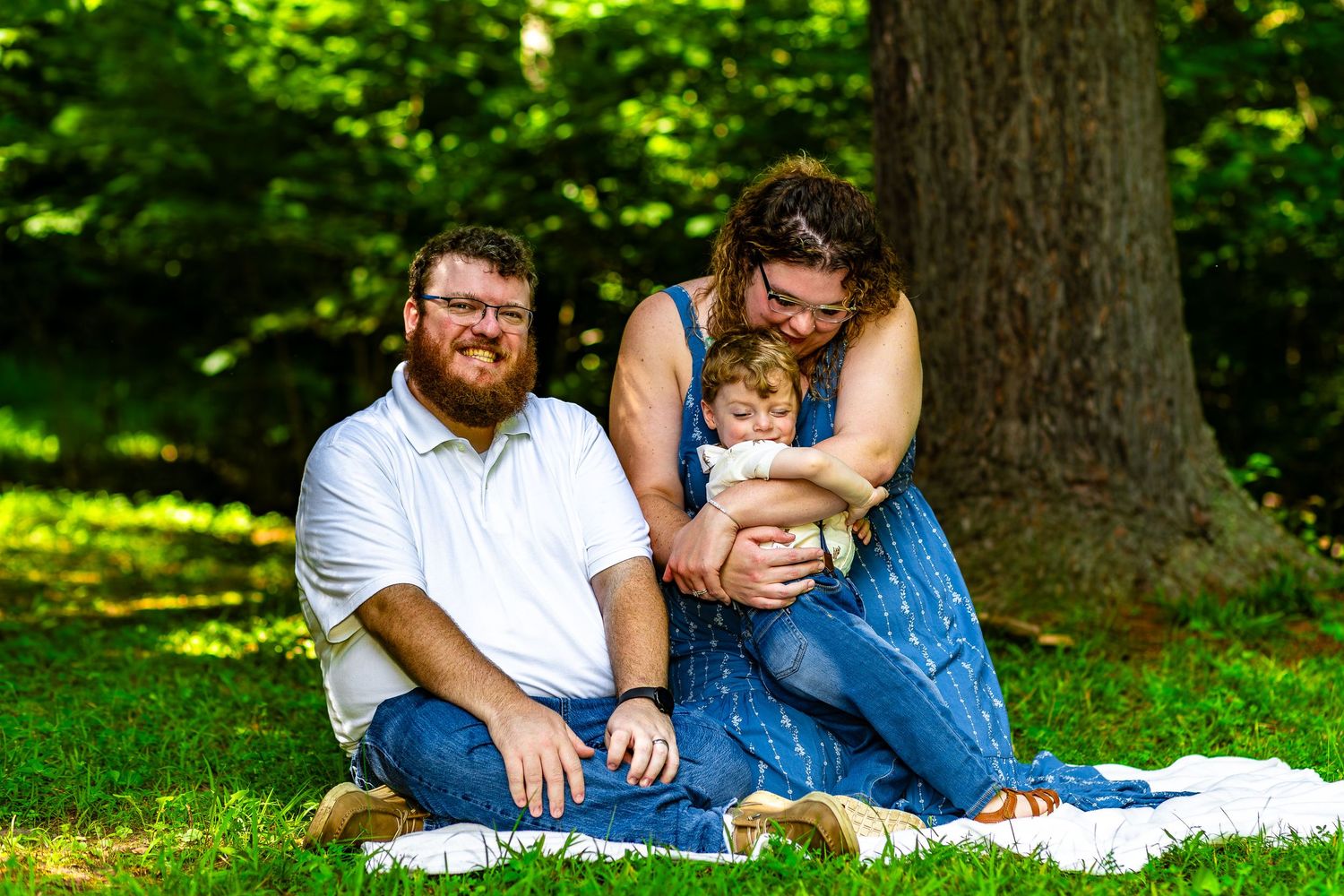 A family sits together on a white blanket enjoying a sunny day in a lush green park setting.