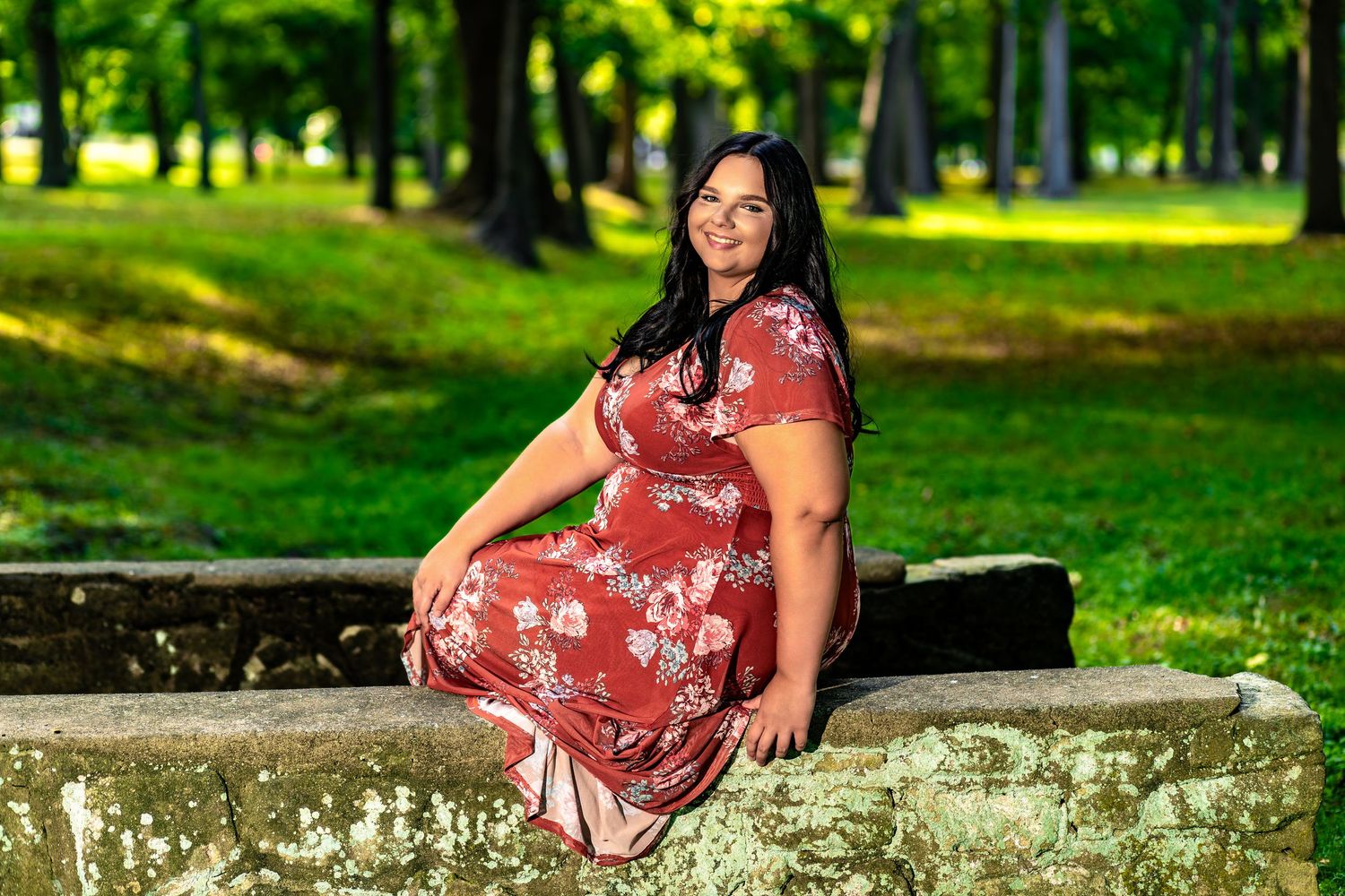 A maternity photo session in a park features someone in a red floral dress sitting on stone steps surrounded by greenery.