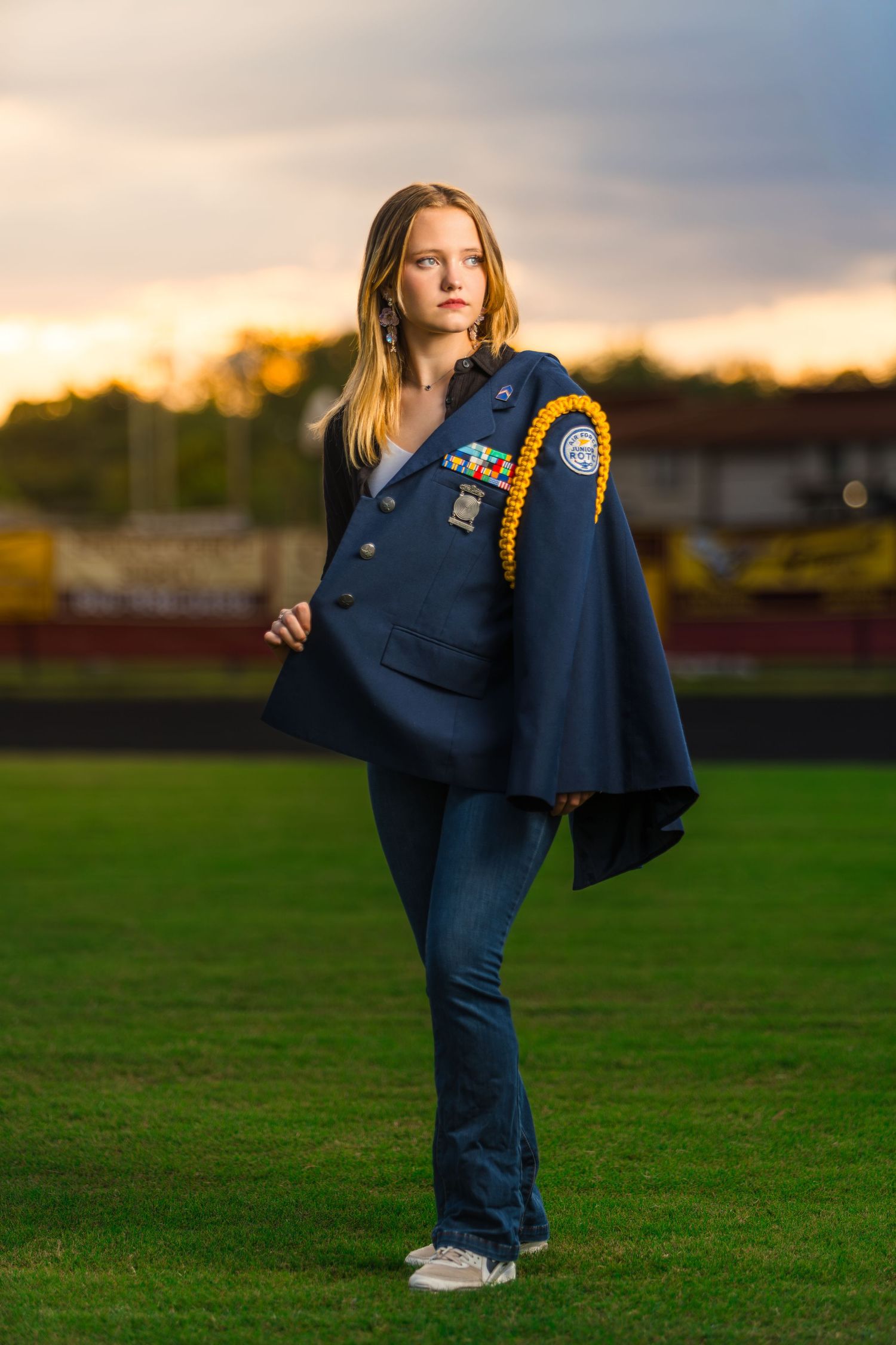 High school varsity student in navy blue uniform poses confidently on green field at sunset.