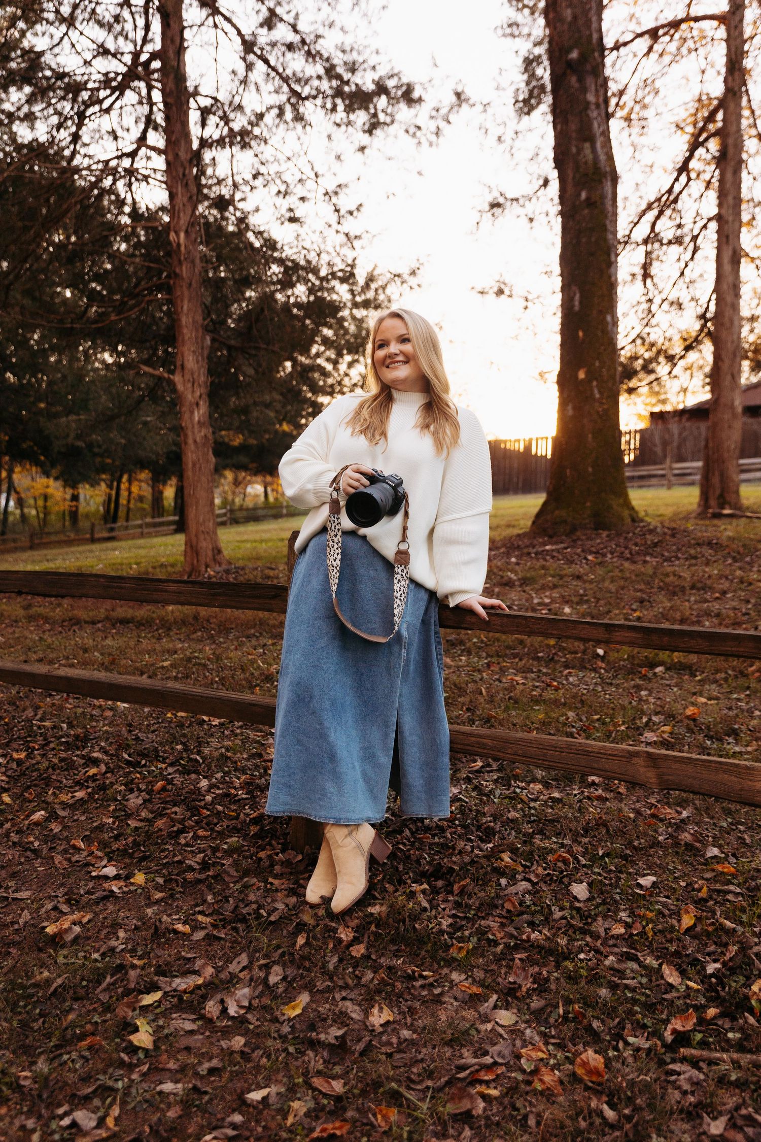 Blonde female photographer smiles holding camera at golden sunset wearing white sweater and denim midi skirt.