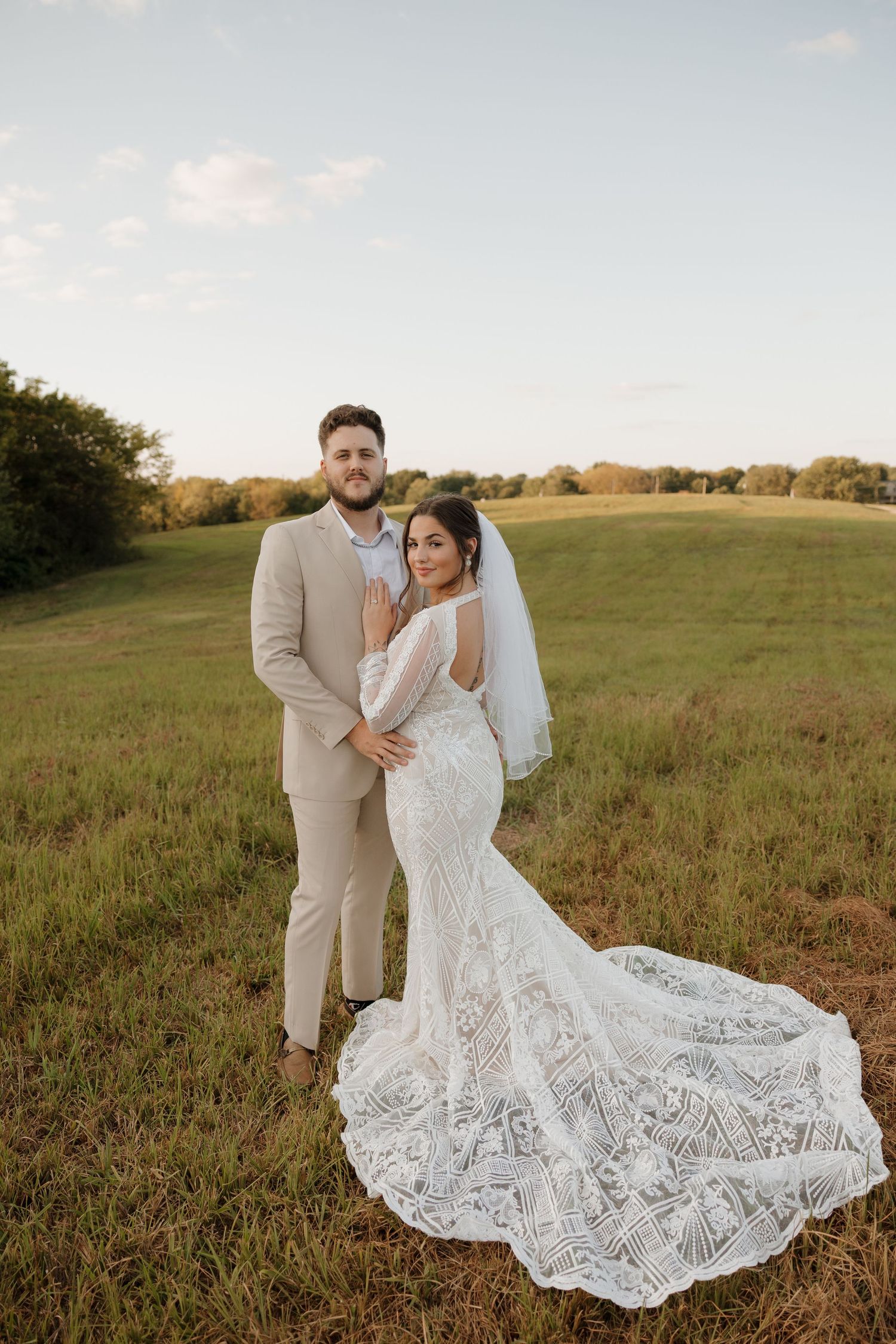 Bride in lace mermaid gown with long train stands with groom in tan suit in green field.