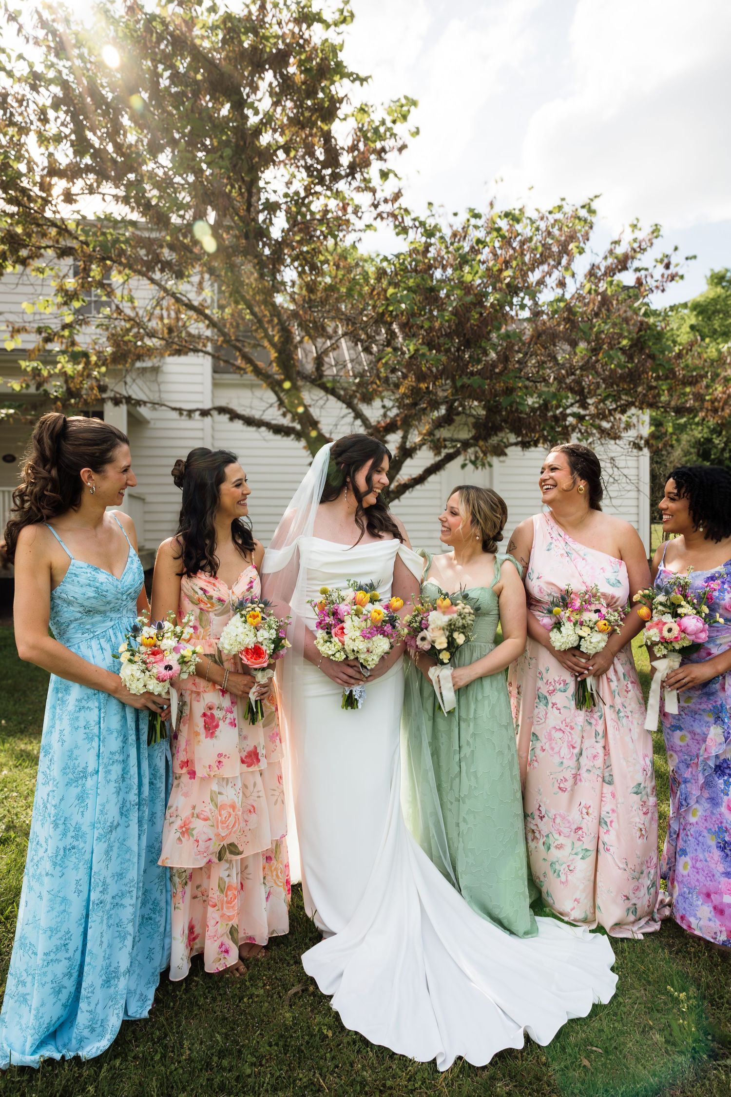 Bride in white gown with five bridesmaids in colorful floral dresses holding bouquets outdoors.