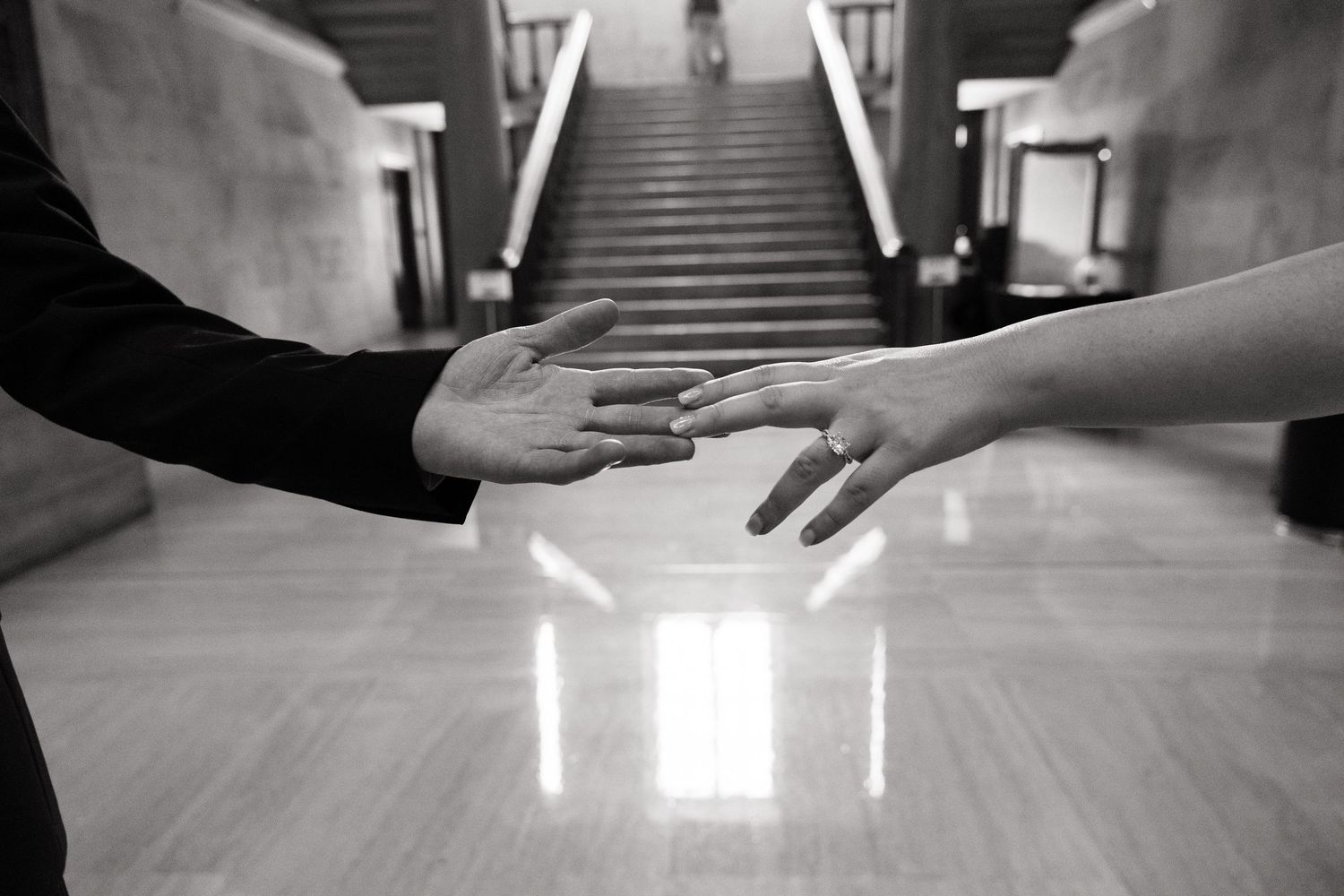 Black and white photo of two people reaching toward each other, hands almost touching, with a grand staircase in the background.