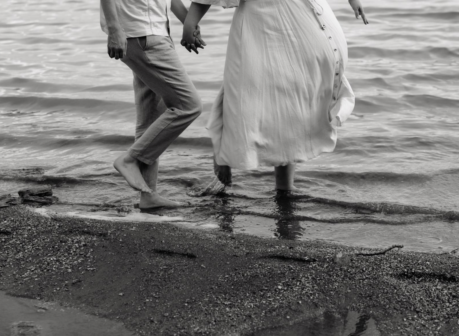 Couple walking barefoot hand in hand along a rocky shoreline in black and white photography.
