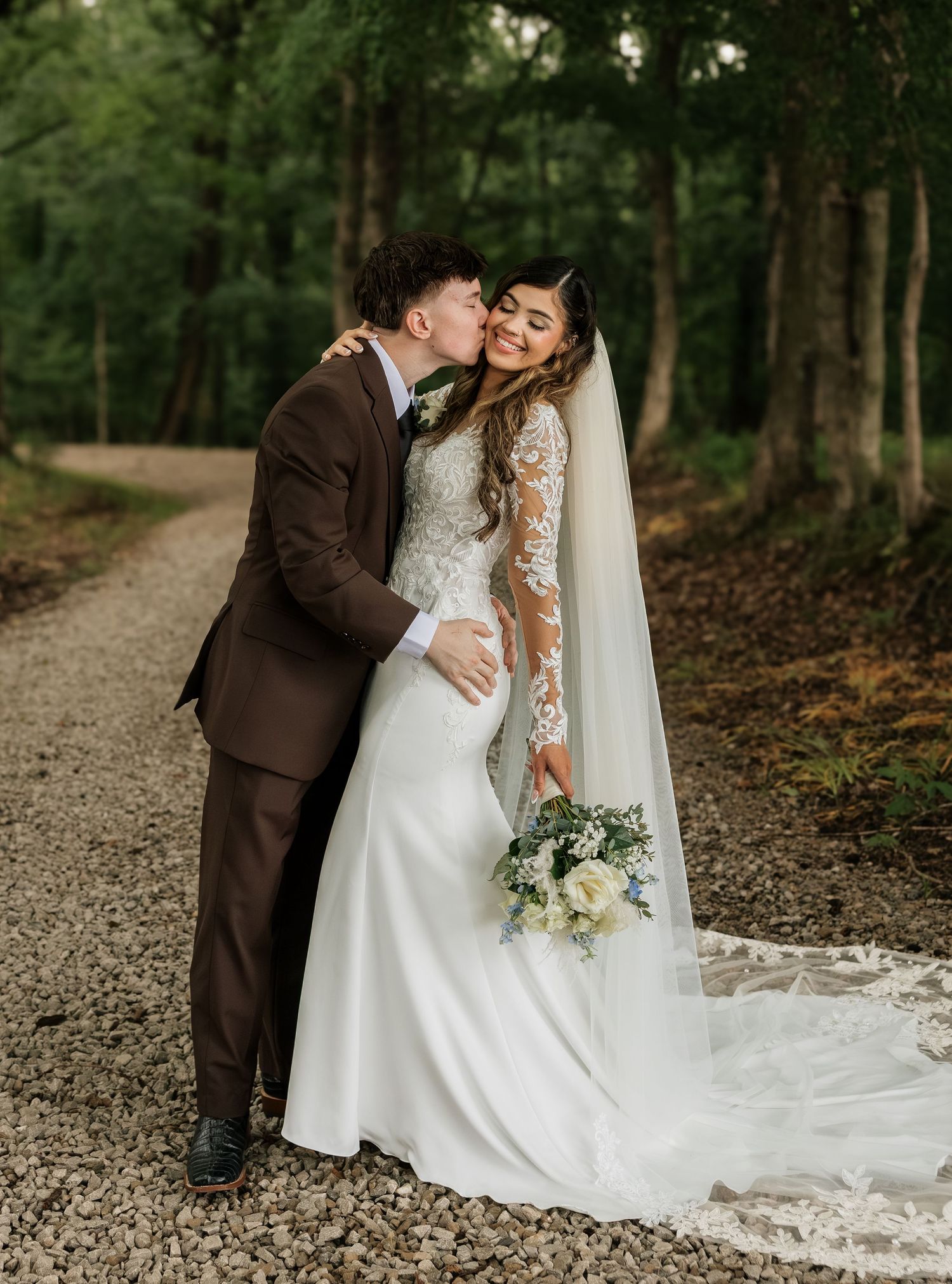 Bride in lace gown with veil holds bouquet as groom in brown suit kisses her cheek on a woodland path.