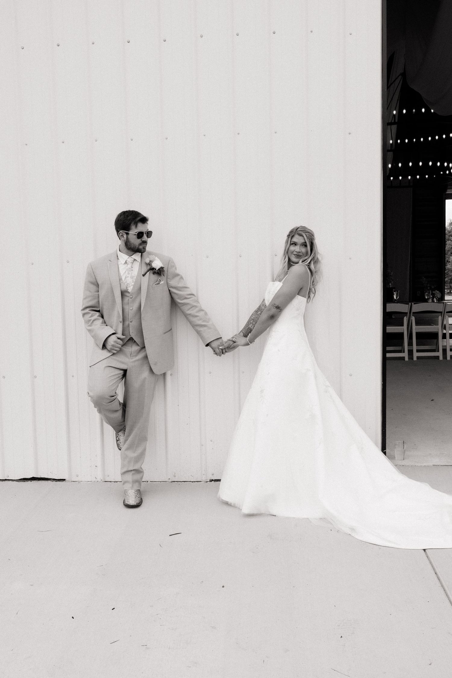Black and white photo of a bride and groom holding hands outside a white barn on their wedding day.